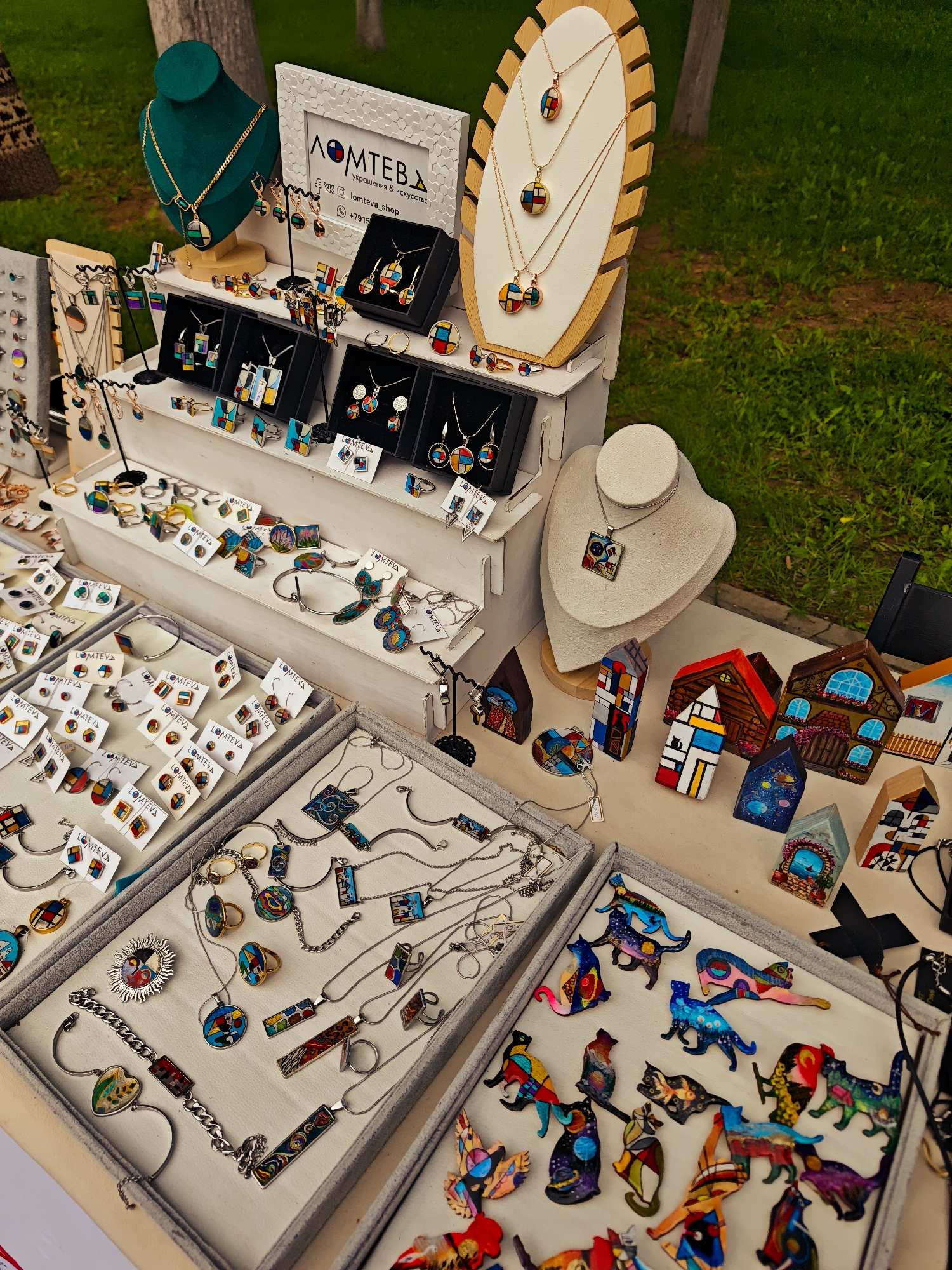 A market display, featuring jewelry and decorative items, is set outdoors. The jewelry, arranged on a multi-tiered display and in trays, includes earrings, necklaces, rings, and bracelets with colorful, geometric designs. Mannequins display necklaces, and a framed sign with a logo is positioned at the back. To the right of the jewelry are decorative house-shaped objects. The background shows greenery and tree trunks.
