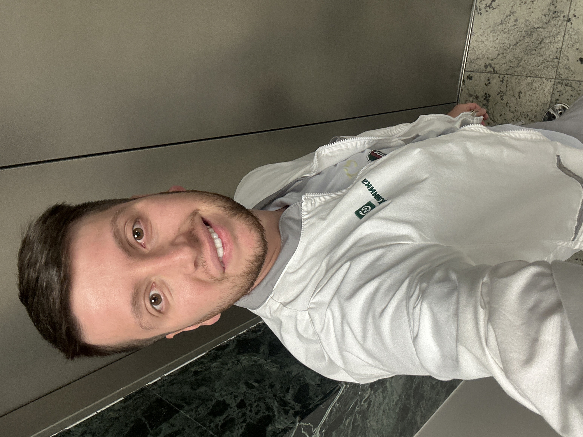 A young adult male, with brown hair, light brown eyes, and light facial hair, is depicted from the chest up, smiling directly into the camera in a selfie. He is wearing a white bomber-style jacket featuring a grey collar and zipper. On the left chest area of the jacket, a green square logo with rounded corners is visible, positioned above the word "КЛИНИКА" (KLINIKA) printed in green Cyrillic script. A light grey garment is visible underneath the jacket. The background on the left side of the frame features a dark green, polished wall with prominent white veining, consistent with marble. On the right side of the frame, a plain, light grey vertical surface is visible, separated by a thin dark vertical line, suggesting an elevator door. The floor, visible at the bottom right, is composed of a light-colored, speckled stone, possibly granite or terrazzo. Based on the Cyrillic text "КЛИНИКА" (Clinic), the location is inferred to be a medical facility within the city of Moscow, Russia.
