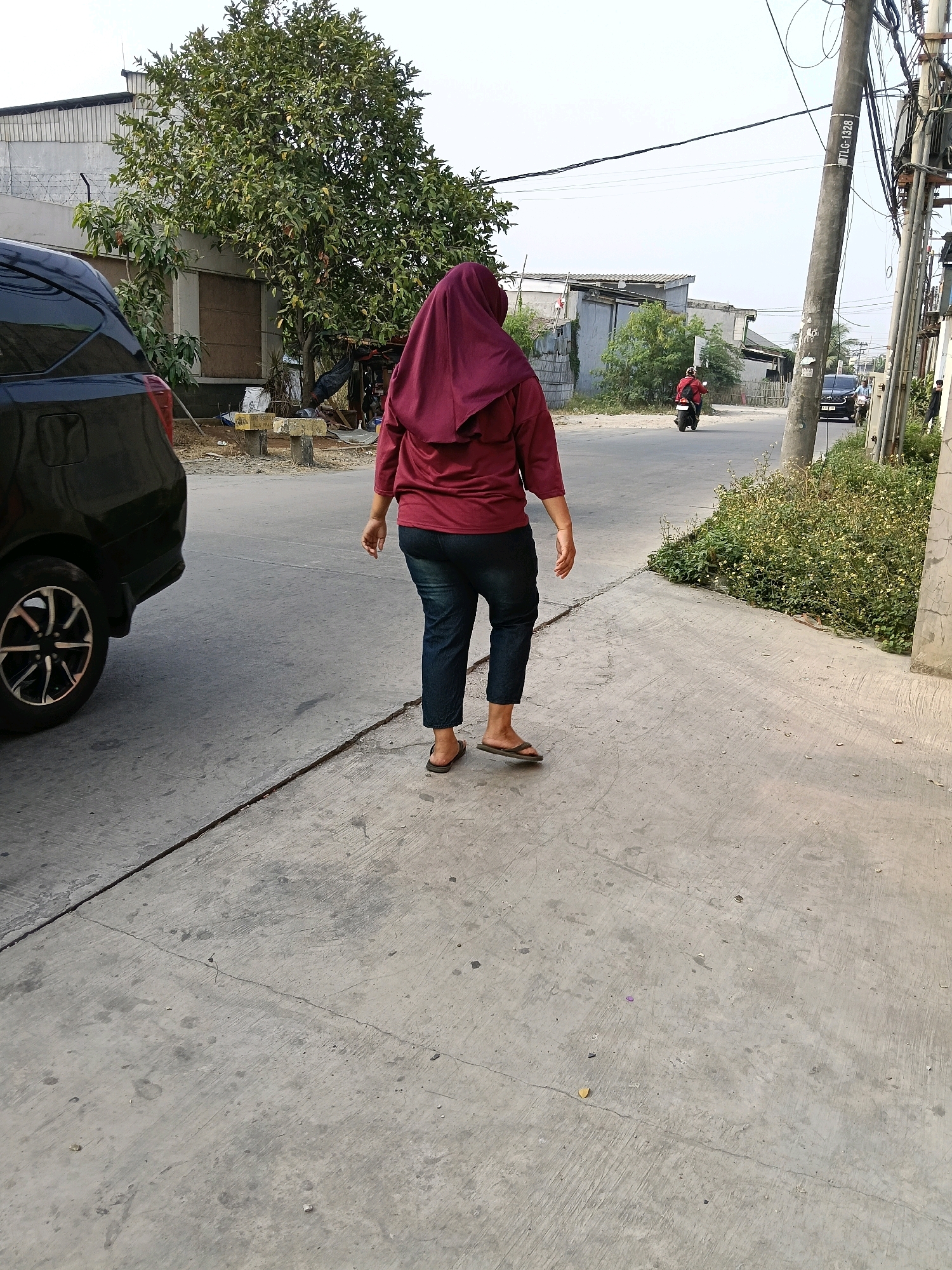 The image depicts a street scene in the city of Belimbing, Indonesia. In the foreground, a person wearing a maroon hijab and dark pants walks away from the camera on a concrete sidewalk. To the left, a black car is partially visible. The background shows a street with buildings, trees, and utility poles. A motorcycle is seen in the distance on the road. The environment appears to be a residential or commercial area.