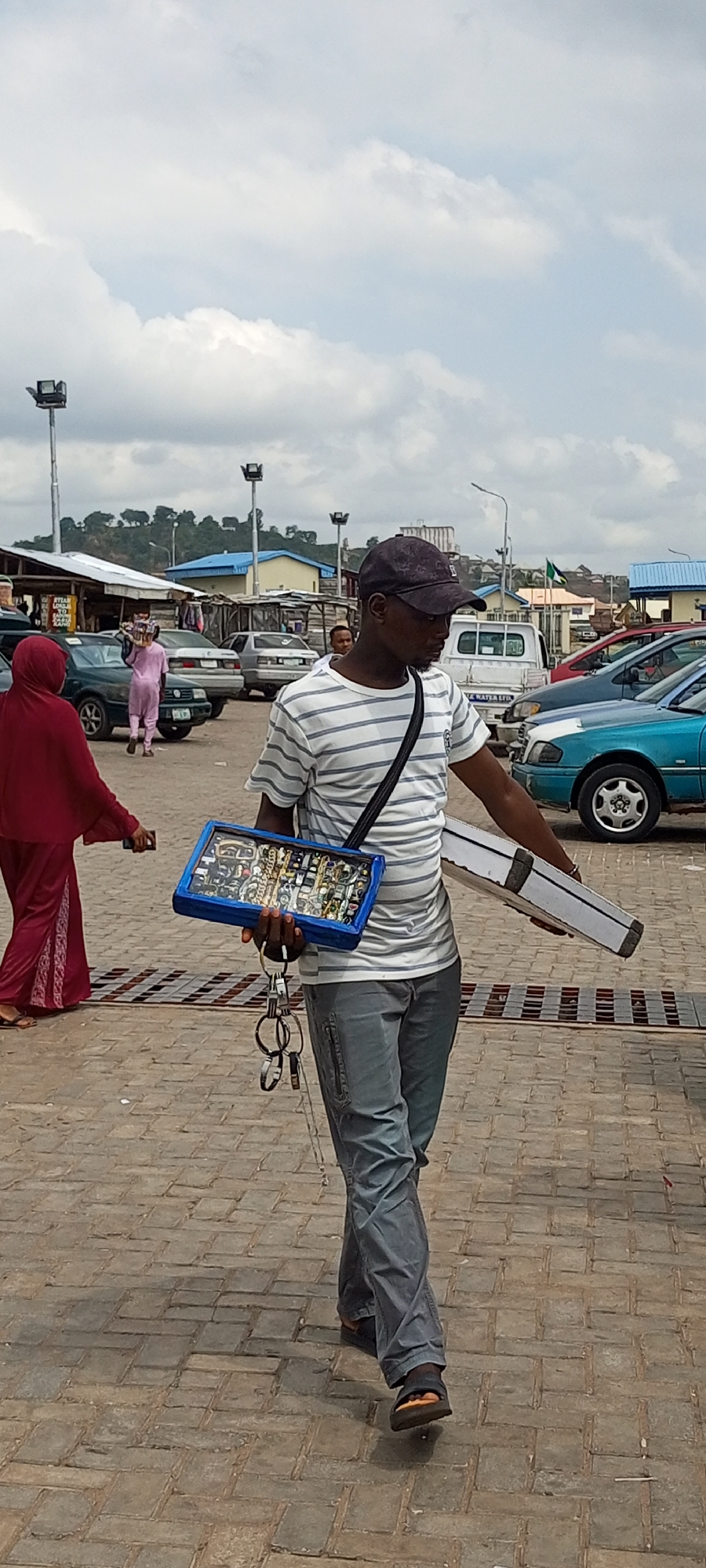 In a bustling outdoor location, likely a market area, in Lokoja, Nigeria, a vendor wearing a striped shirt, grey trousers, and a dark cap walks across a brick-paved area. He has a black strap across his shoulder, carrying a blue display case filled with jewelry in one hand and a white rectangular case in the other. Dangling from his hand are several keys on a ring. A woman in a red head covering and robe walks in the opposite direction to the left of the vendor. Several cars are parked nearby, and in the background, there are buildings, street lights, and a cloudy sky.