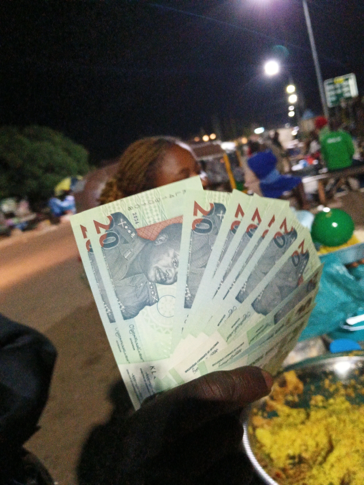 A close-up, eye-level shot shows a hand fanning out several 20-unit currency notes, appearing to be Nigerian Naira, in Bukuru, Nigeria. The background is dark and slightly out of focus, suggesting it is nighttime. A street lamp with a bright light source is visible in the upper center of the frame, casting a glow. Various blurred figures and objects are present in the background, indicating a public outdoor setting, possibly a market or roadside area. To the right, a green spherical object rests on a turquoise cloth, near a metallic bowl filled with yellow, granular food. The currency notes are light green with images of individuals printed on them. The overall impression is a casual, nighttime scene focused on the display of cash and food.