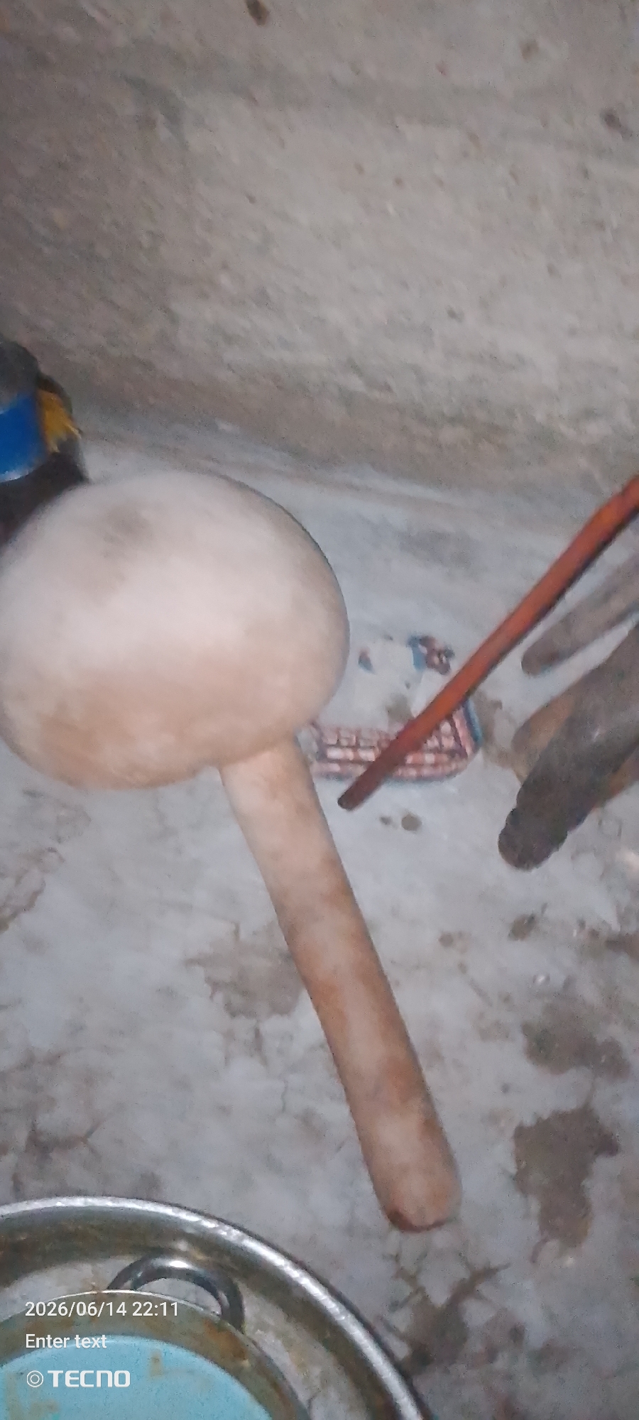 A rustic scene unfolds in Jalingo, Nigeria, with the primary focus on a large, light-brown, gourd-like object resting on a dusty, concrete floor. The object, resembling a traditional pestle or a decorative item, has a spherical top and a cylindrical handle. Its surface appears worn and textured. To the right of the gourd, a dark brown, rough-textured object, possibly a piece of wood or a root, lies on the floor. Between these two objects, a colorful textile with a repeating pattern, possibly a mat or fabric, is partially visible. In the upper right quadrant, a thin, reddish-brown stick is diagonally positioned. In the lower left corner, a metallic bowl or container with visible handles is present, with a hint of light blue inside. The background is a textured, light-colored wall, likely made of concrete or plastered earth, showing signs of age and wear. The overall impression is of an indoor space with a simple, utilitarian ambiance. The lighting is dim, casting subtle shadows and highlighting the textures of the objects. The presence of the Tecno logo and a date stamp suggests the image was captured by a mobile phone.