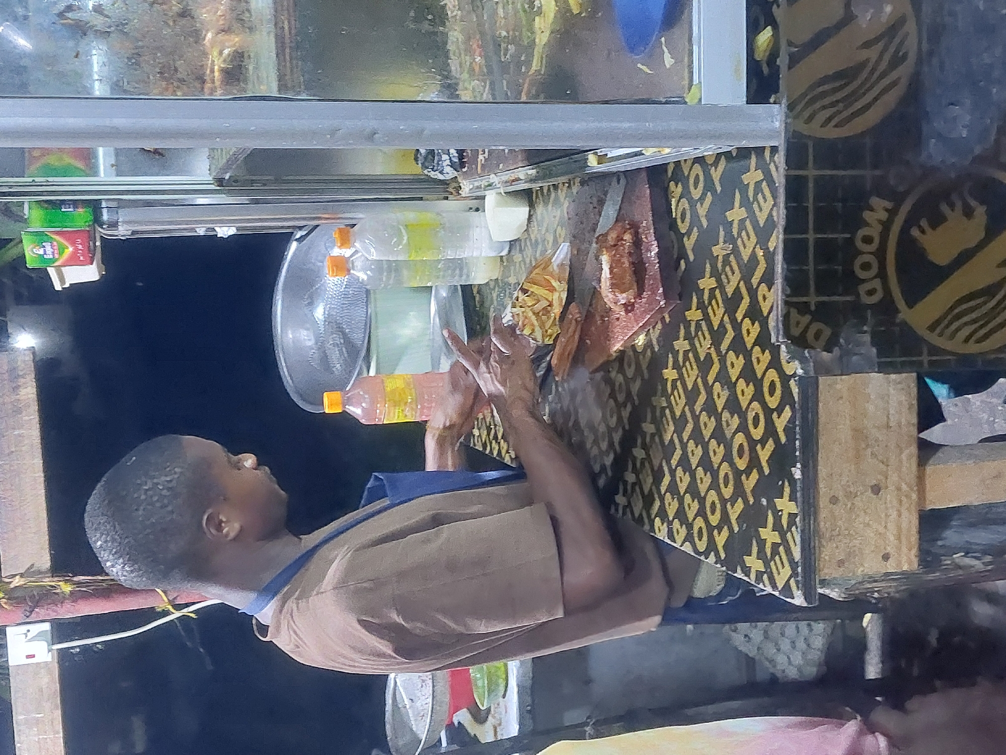 A street vendor in Dar es-Salaam, Tanzania, prepares food on a food cart. He wears a brown uniform with a blue apron. On the cart, food like fries and meat are arranged on a cutting board. Bottles of liquids and food containers are also present. The cart is made of metal and wood with branding logos visible. The background shows a night scene in the city.