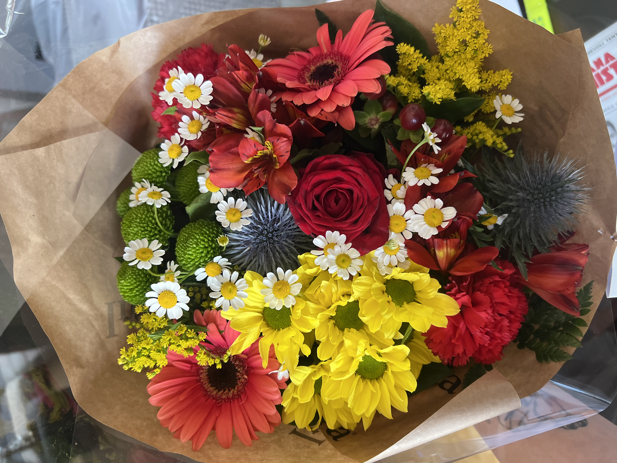 A vibrant bouquet of mixed flowers is presented wrapped in brown paper. The flowers include red and orange Gerbera daisies, a red rose, yellow chrysanthemums, small white daisies, green chrysanthemums, red Alstroemeria, yellow solidago, berries, and a blue thistle. The bouquet sits on a reflective surface under artificial light. A book is visible in the background. There is no evidence to confirm a location in Milan, Italy.