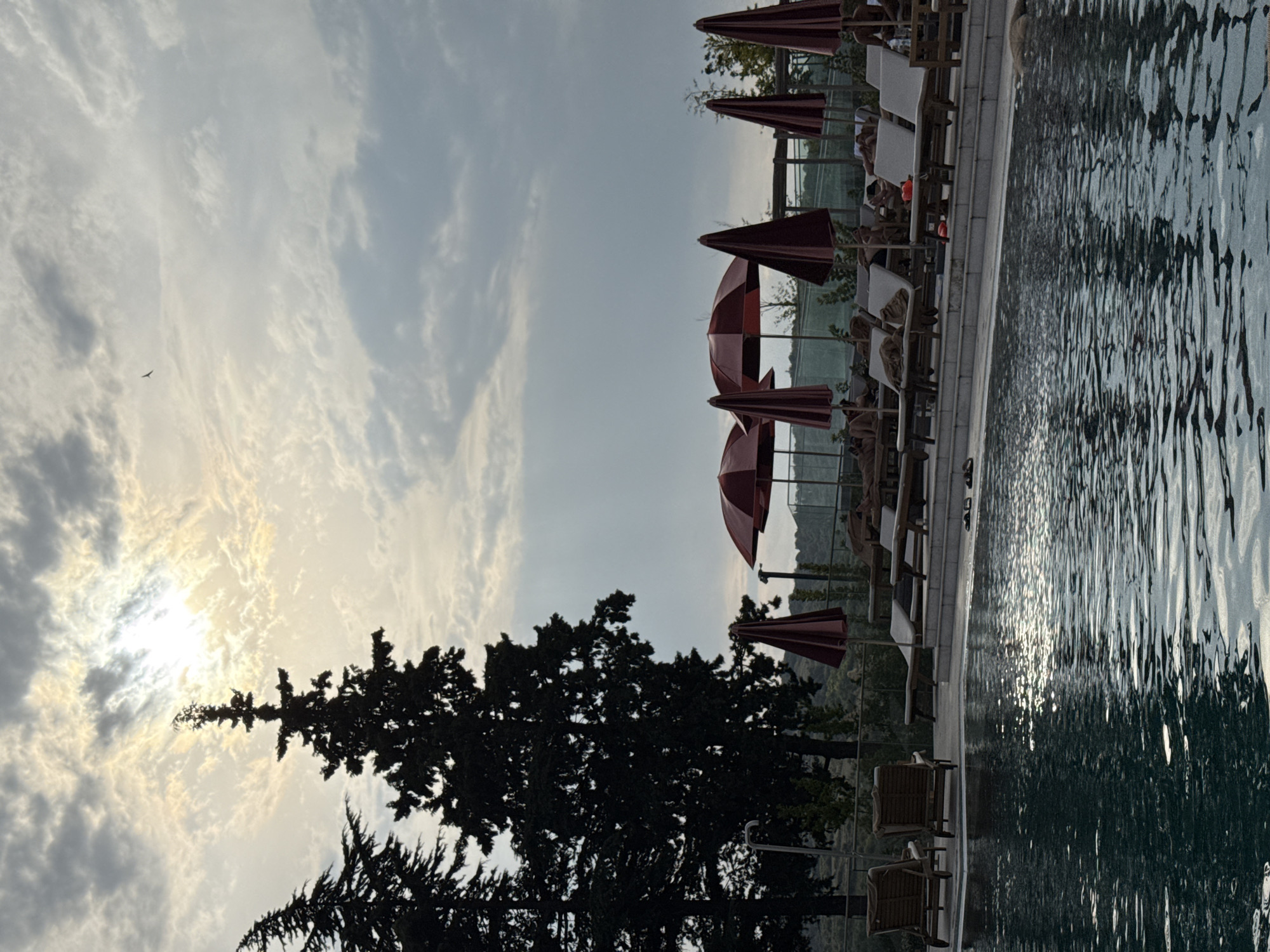 This image captures a serene outdoor scene, likely at a resort or hotel in Sighnaghi, Georgia. In the foreground, a swimming pool reflects the overcast sky, with sunlight peeking through the clouds, creating a dappled effect on the water's surface. Several lounge chairs with red umbrellas are positioned along the poolside, some occupied by individuals relaxing. Trees frame the left side of the image, with their dark silhouettes contrasting against the bright sky. In the distance, hills and vegetation suggest a rural or hilly landscape, typical of the Kakheti region. The overall atmosphere is peaceful and tranquil, evoking a sense of relaxation and leisure.