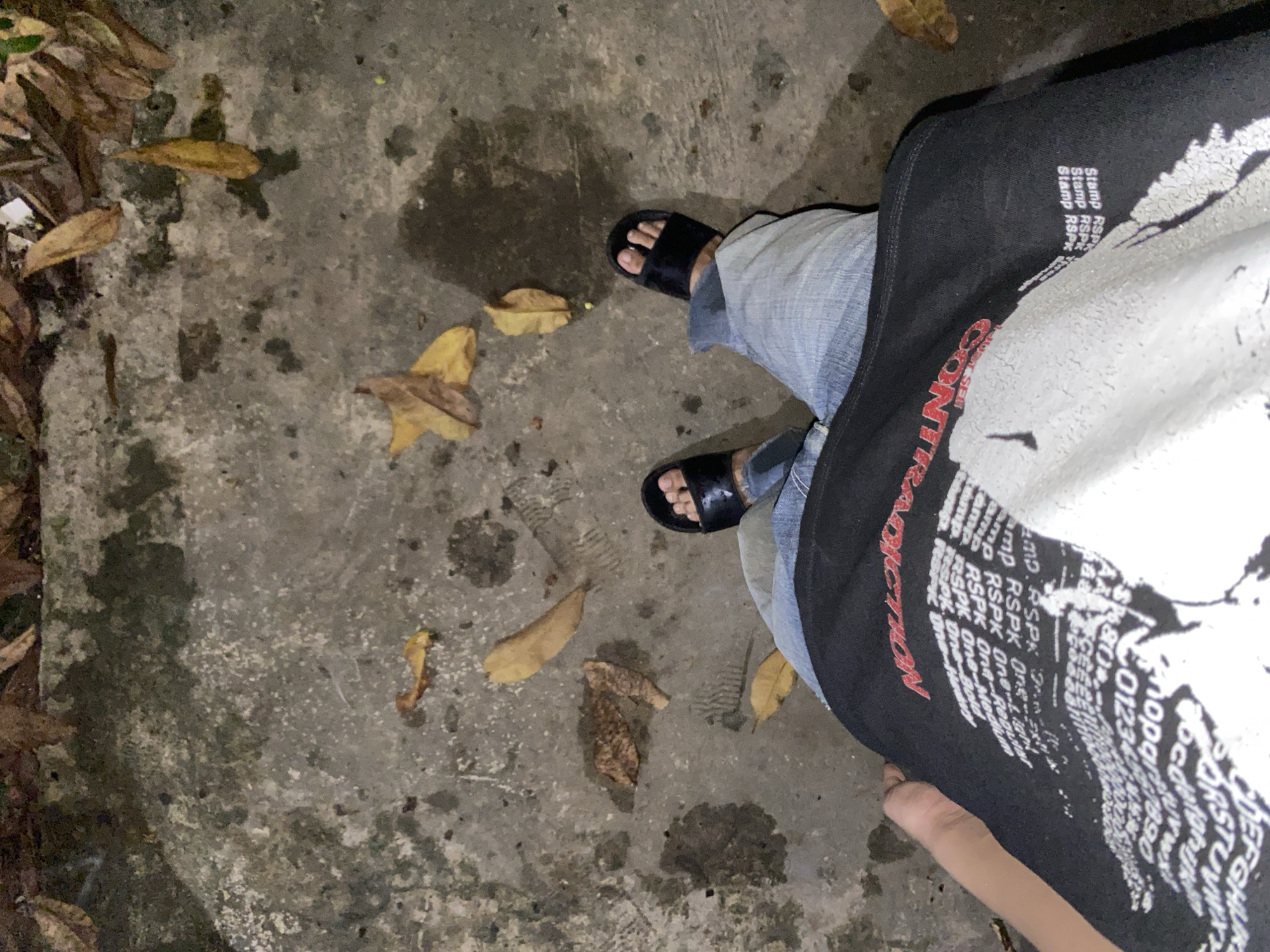 A first-person perspective image shows a person standing on a wet, concrete surface covered in fallen leaves. The person is wearing black slides, blue jeans, and a black t-shirt with white and red text and graphics. The t-shirt has the word "CONTRADICTION" printed in red across the chest. A blurred portion of the person's arm is visible on the left side of the frame. The environment appears to be outdoors, possibly a sidewalk or pathway, with scattered brown and yellow leaves. The concrete shows signs of dampness and some staining. No specific landmarks indicating Banjarmasin, Indonesia are visible.