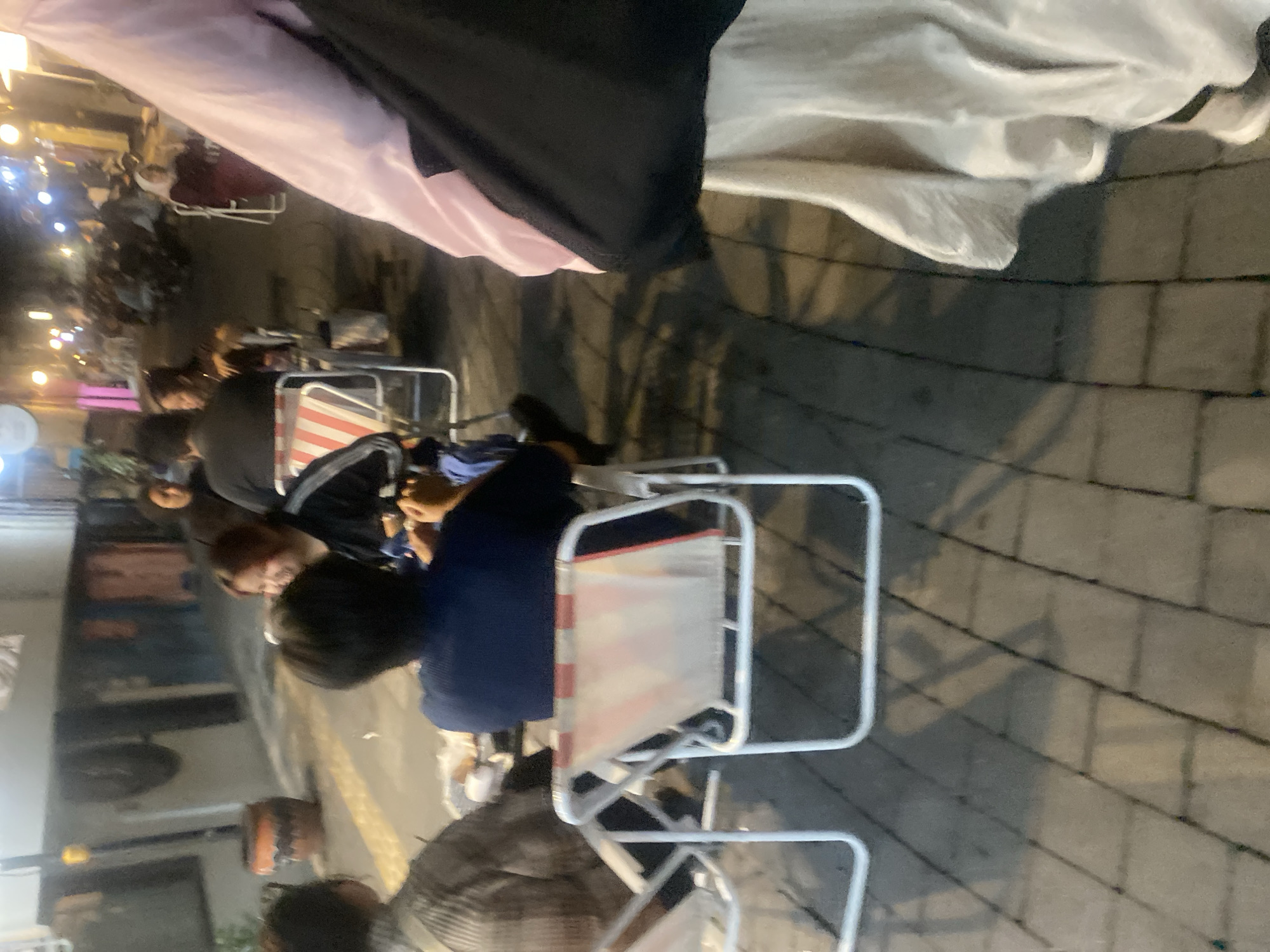The image depicts a nighttime scene on a pedestrian walkway paved with light-colored bricks, likely in Banjarmasin, Indonesia. Several people are seated at tables, suggesting an outdoor dining or gathering area. Two individuals are visible in the foreground: one is wearing a blue shirt and seated in a white folding chair with red and white striped fabric. Another person in a plaid shirt is partially visible to the left, also seated. Further back, a woman with short, dark hair and a black jacket is visible. In the middle ground, a woman wearing a maroon hijab and dress is seated in a white folding chair. Various lights illuminate the area, with some visible in the background indicating buildings and possibly a street. A large, reddish-brown decorative pot is present on the left side of the walkway. The overall atmosphere suggests a casual, communal setting.