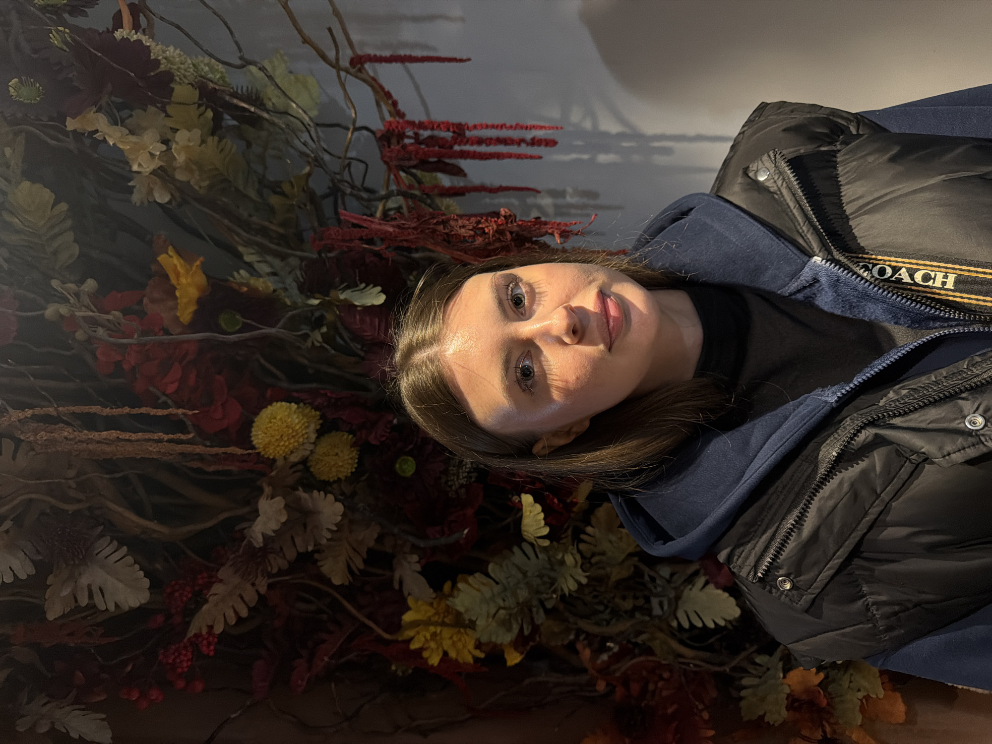 The image shows a woman with light brown hair and fair skin posing against a backdrop of dried flowers and branches. She is wearing a dark blue hoodie and a black puffer jacket with "COACH" printed on the zipper trim. The background consists of various dried flowers in shades of red, yellow, and white, creating a textured and somewhat autumnal scene. The woman's face is well-lit. The image appears to have been taken indoors with artificial lighting. There is no information in the image that would indicate the location as Chaikovsky, Russia.