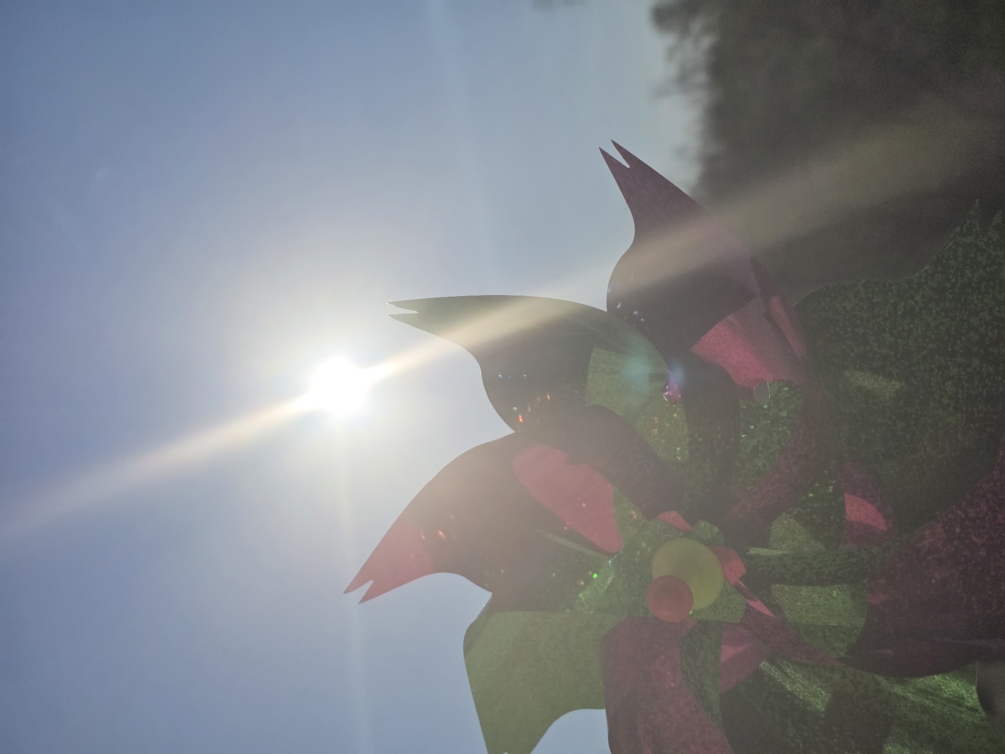 A close-up low-angle shot of a multi-colored pinwheel against a bright blue sky with the sun shining directly in the background, making the pinwheel partially silhouetted. The pinwheel's blades are a mix of red and green, with what appears to be glitter on the surface. The center of the pinwheel features red and yellow elements. The sun is positioned to the left of the pinwheel, creating a lens flare effect. The environment includes a blue sky and the possible edge of some greenery.
