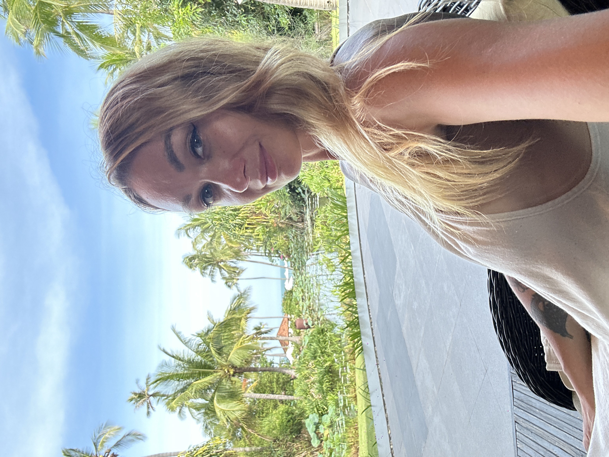 A blonde woman with blue eyes is captured in a close-up selfie. She is looking towards the camera with a slight smile and her hair is falling over her shoulder. She is wearing a light-colored top. The background suggests a tropical location, with lush green palm trees, dense foliage, and a body of water with lily pads. A hint of a tiled patio is visible in the foreground. The image appears to be taken outdoors.