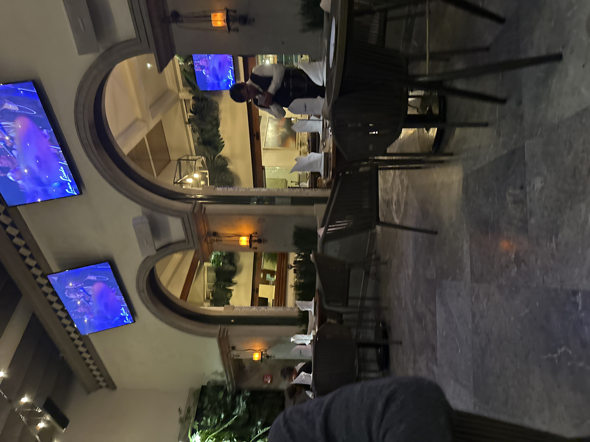 The image depicts a restaurant in Mexico City, Mexico, with an outdoor seating area under arched openings. A waiter dressed in a white shirt and dark vest is attending to a table. Modern outdoor furniture is arranged for dining, and televisions mounted on the walls display colorful visuals. Greenery is visible in the background, adding to the ambiance.