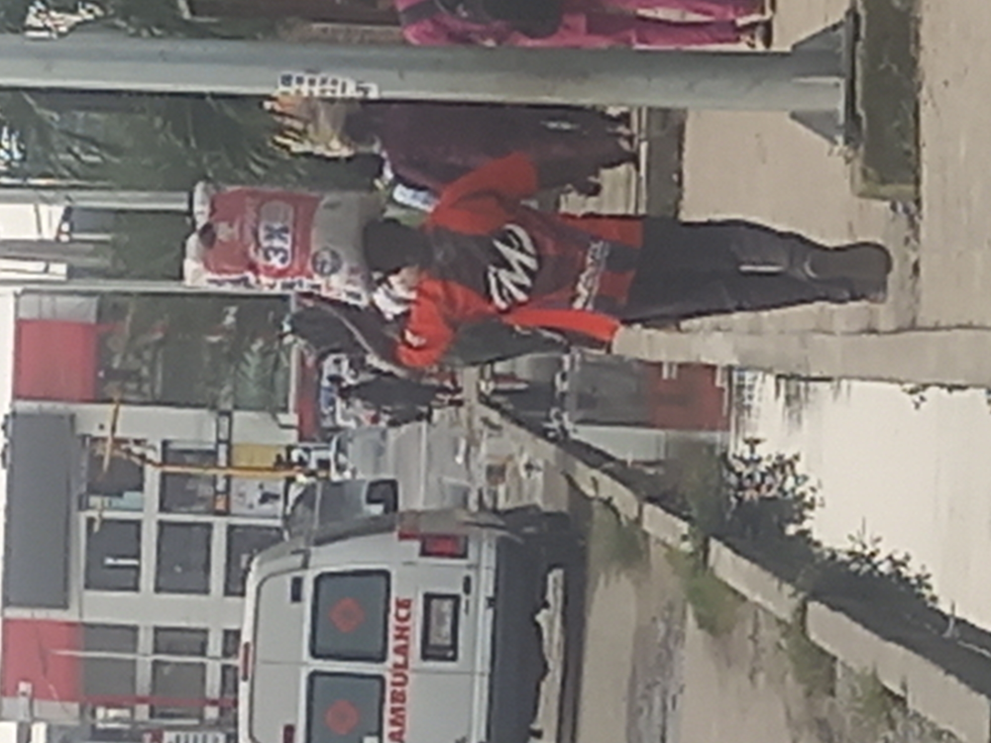 The image shows a street scene in Ilado, Nigeria. In the foreground, a person is walking from right to left, carrying a large white bag with red lettering on their head. The person is wearing a red and black long-sleeved shirt and dark pants. To the left of this person, an ambulance is parked. The ambulance is white with "AMBULANCE" written in red on the side. Further to the left, a multi-story building with white and red sections is visible. The street has a concrete drainage ditch running alongside the sidewalk. Other people and vehicles are present in the background, suggesting a busy urban environment. Palm trees are visible in the background, indicating a tropical climate. The overall impression is that of a typical street scene in a Nigerian city.