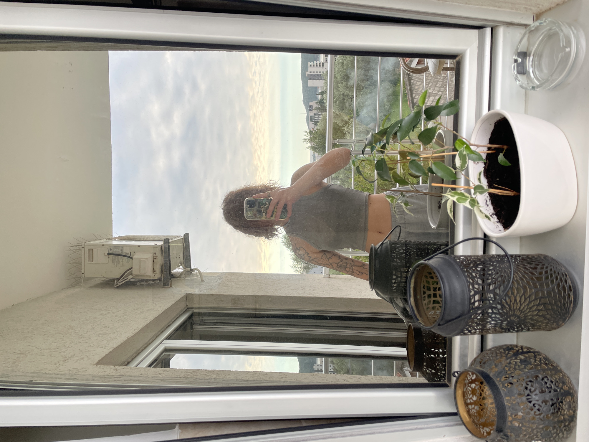 The image shows a person taking a selfie in a mirror. The reflection shows a woman with curly hair, a gray tank top, and tattoos on her arm, holding a phone. Behind her is a view of Rafailovići, Montenegro, with buildings, trees, and a cloudy sky. On the window sill in front of the mirror, there is a white potted plant and several decorative metal lanterns. An air conditioning unit is visible on the exterior wall outside the window.