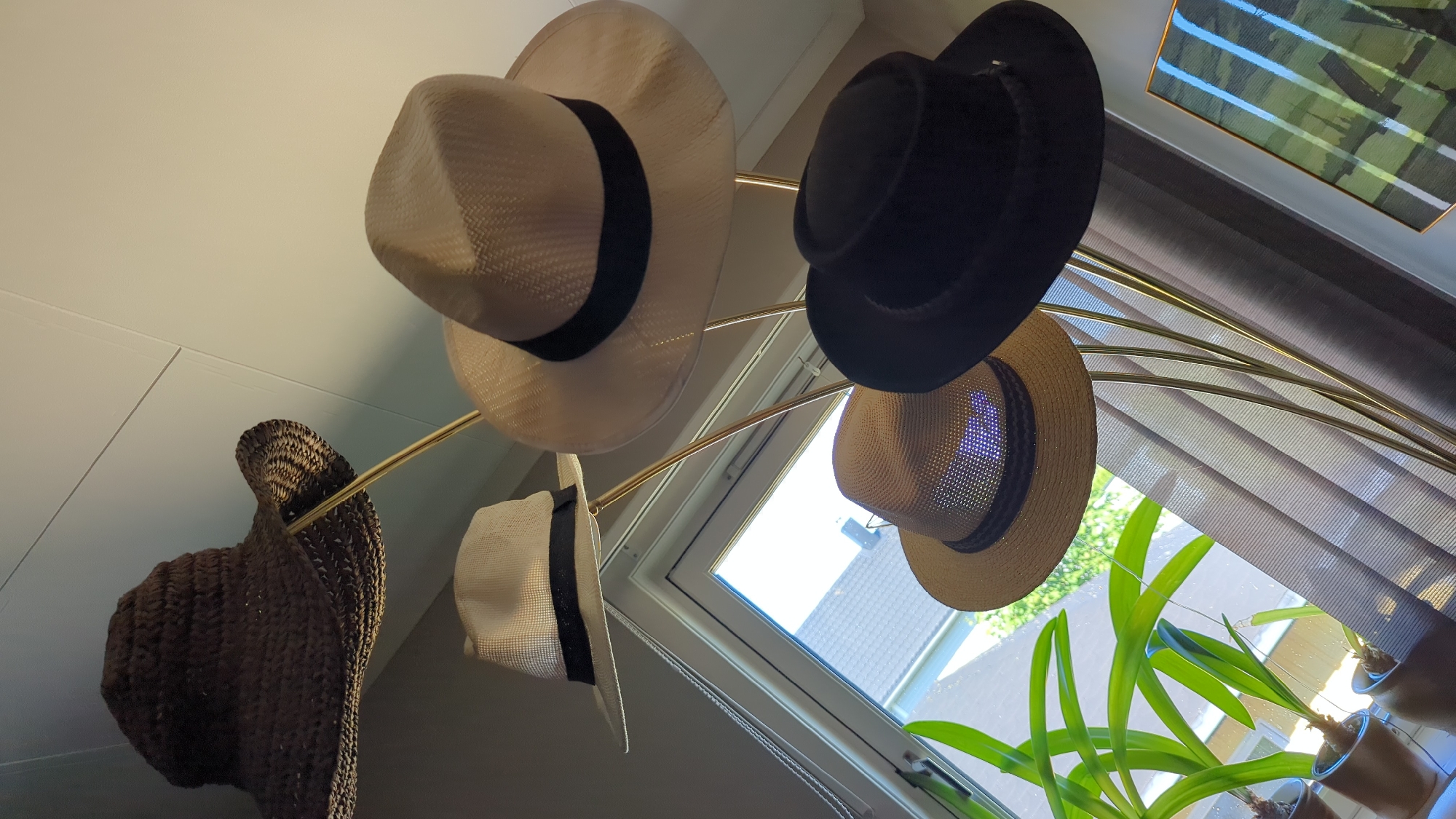The image displays a collection of hats on display. A dark, wide-brimmed crocheted hat is positioned at the top left, with a lighter-colored fedora-style hat with a black band below it. To the right of the fedora is another light-colored, woven hat with a black band. Below these, a straw hat with a darker band is visible, and to its right, a dark-colored, possibly felt hat. The hats are arranged on a multi-tiered golden stand. In the background, through a window, residential buildings and greenery can be seen. Curtains are drawn on the right side of the window, and a potted plant with long green leaves is situated on the windowsill on the left. A framed artwork with green and blue stripes hangs on the wall to the right. The lighting suggests an indoor setting, likely a room in Kløfta, Norway, with natural light coming from the window. There are no people visible in the image, and no actions or interactions are depicted. The scene is static and focuses solely on the arrangement of the hats and the surrounding environment.