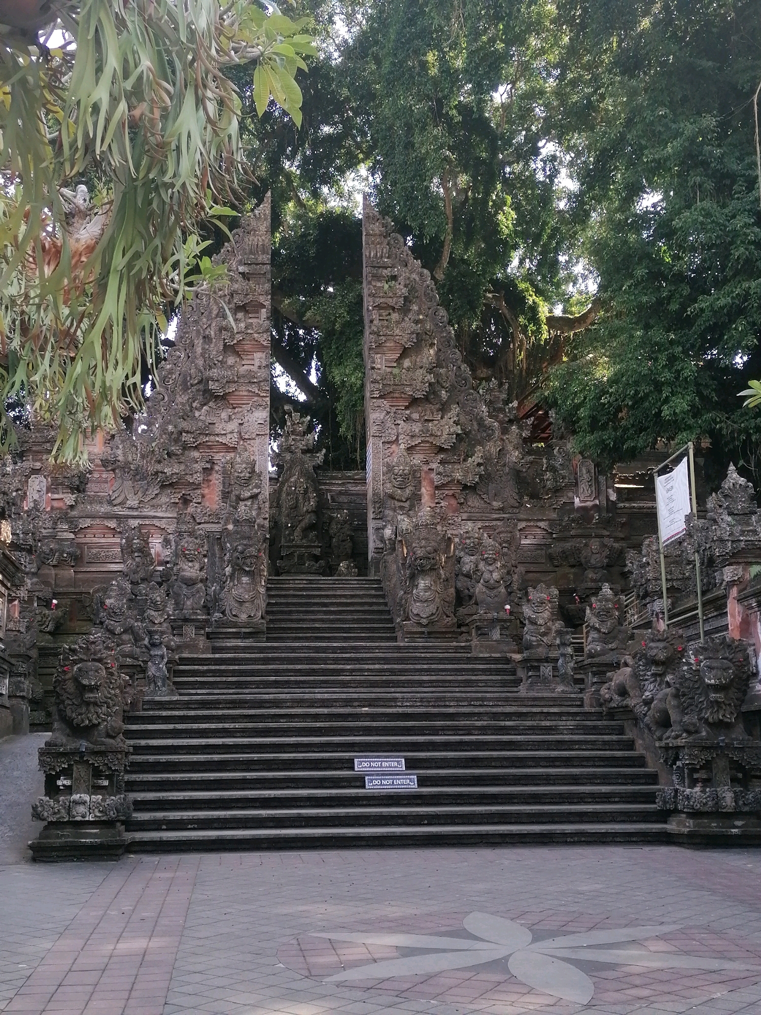 This is a full, eye-level, medium shot photograph of a large, stone temple gate and a wide set of stairs leading up to it. The temple architecture is ornate and features many detailed carvings of figures and mythical creatures. Flanking the stairs on either side are large, stone statues of lions. The gate is set within a dense, green forest, with a large tree visible behind the gate and branches of a lighter green plant hanging in the foreground from the left side of the frame. The ground in front of the stairs is paved with square tiles in a light gray and brown pattern, with a faint, light gray flower-like design in the center. Two white signs with red lettering are visible on the stairs. The scene is lit by natural daylight, casting some shadows. The location is identified as Ubud District, Indonesia.