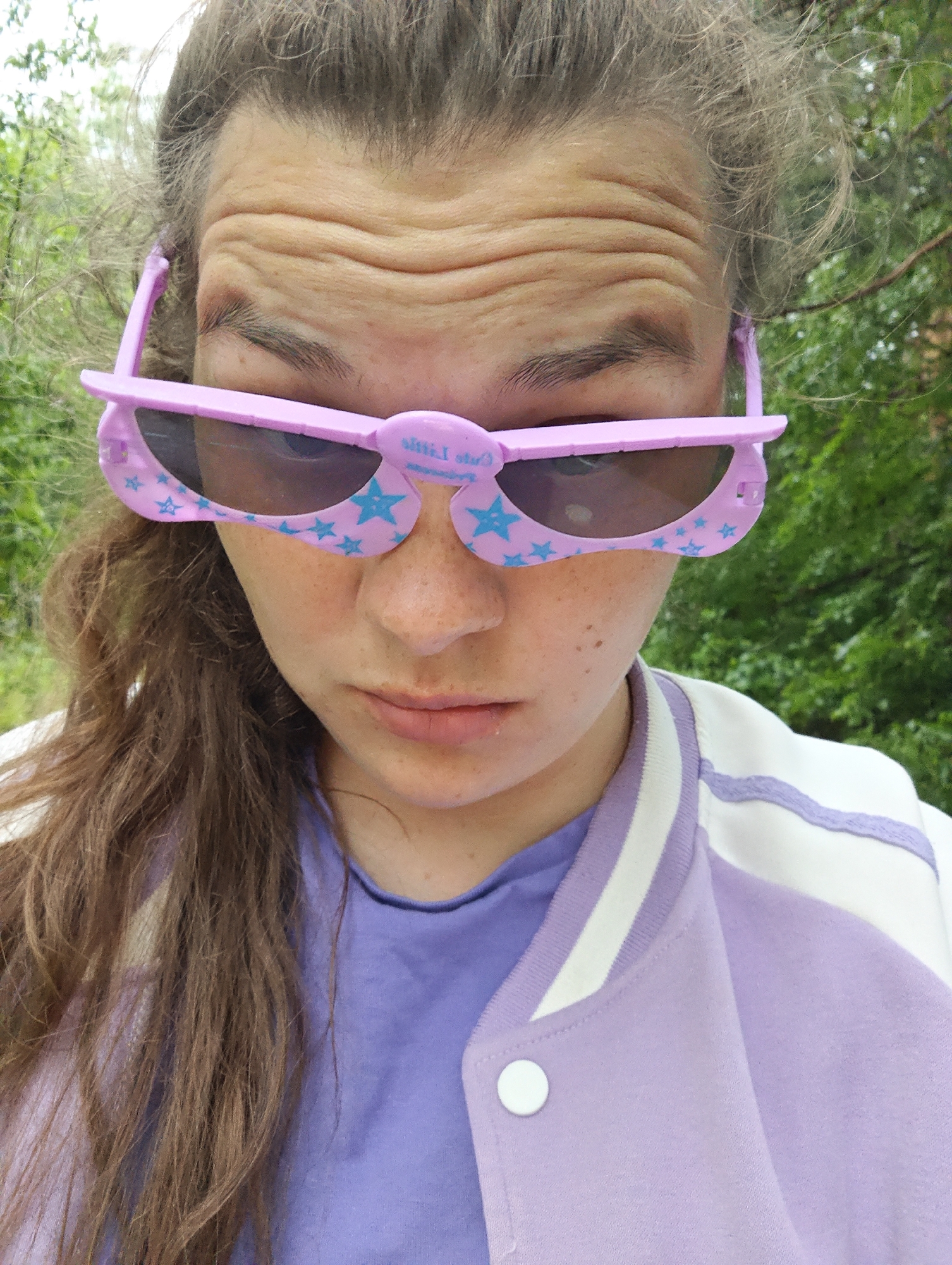 A close-up, eye-level portrait features a young person in an outdoor setting in Chaikovsky, Russia. The subject has long, wavy brown hair and visible freckles across their face. They are wearing distinctive pink novelty sunglasses adorned with light blue stars, which appear to be folded at the bridge, giving them an unusual, raised appearance. A partial text, possibly "Oh Little," is visible on the upper left frame of the sunglasses.
The person's forehead is deeply furrowed, suggesting concentration, discomfort from light, or a specific expression. Their mouth is slightly downturned, contributing to an overall ambiguous expression that could be serious, pensive, or slightly uncomfortable. They are dressed in a purple t-shirt, layered under a white and lavender varsity-style jacket with white stripes on the collar and shoulders, and a white snap button visible on the front.
The background is softly blurred, showing lush green foliage and trees, indicating a natural, outdoor environment like a park or wooded area. The lighting suggests it is daytime, possibly overcast or the subject is in a shaded spot, as there are no harsh shadows. The image captures a moment of posing or a selfie, with the subject's gaze directed towards the viewer.