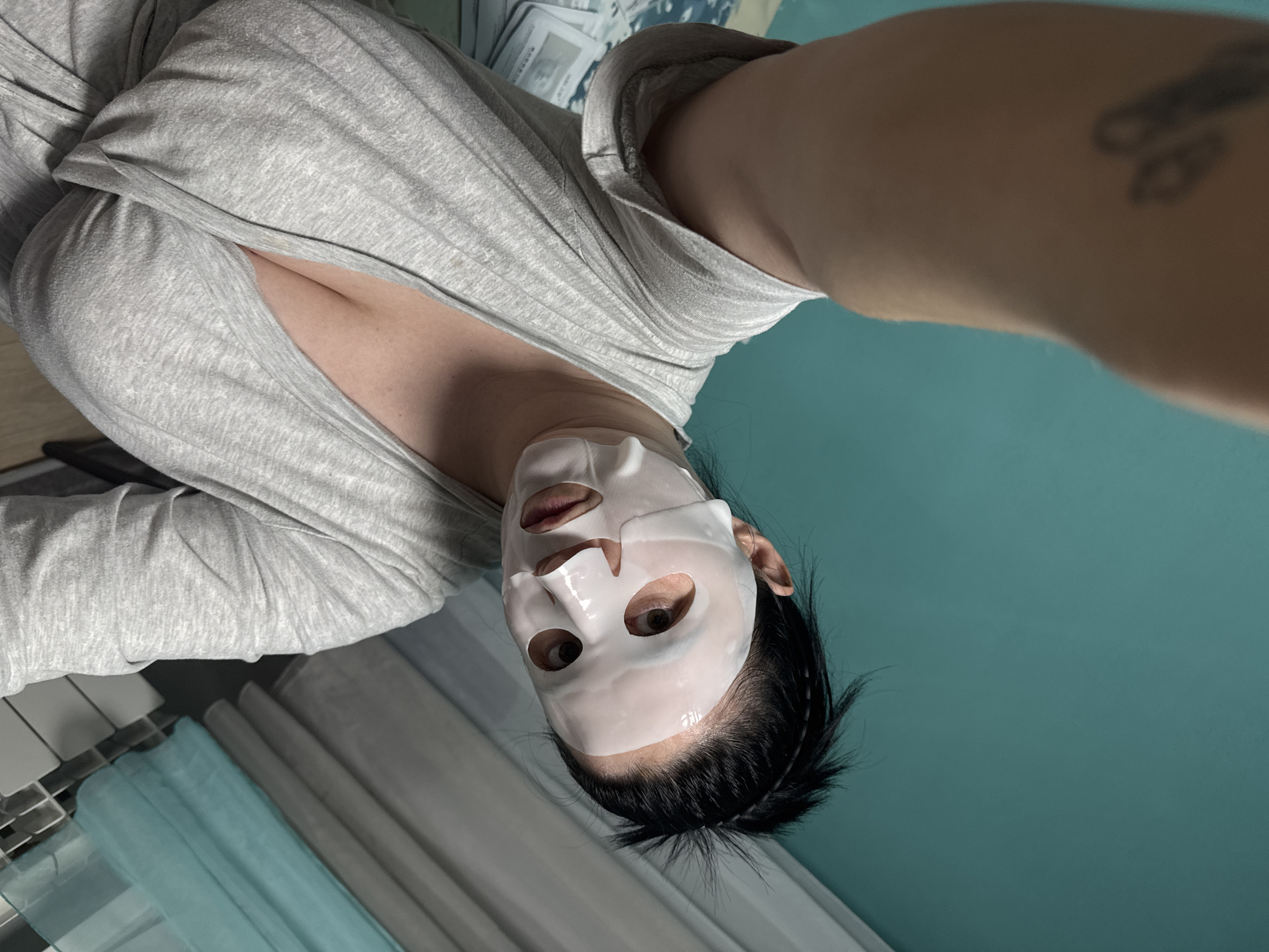 A selfie-style, eye-level, close-up shot features a person wearing a white sheet mask. The person is wearing a light gray, V-neck shirt. A tattoo is visible on the person's right arm. The background includes a teal-colored wall and, in the lower left corner, what appears to be a radiator and some fabric. The location, Zav'yalovo, Russia, is not determinable from the image.