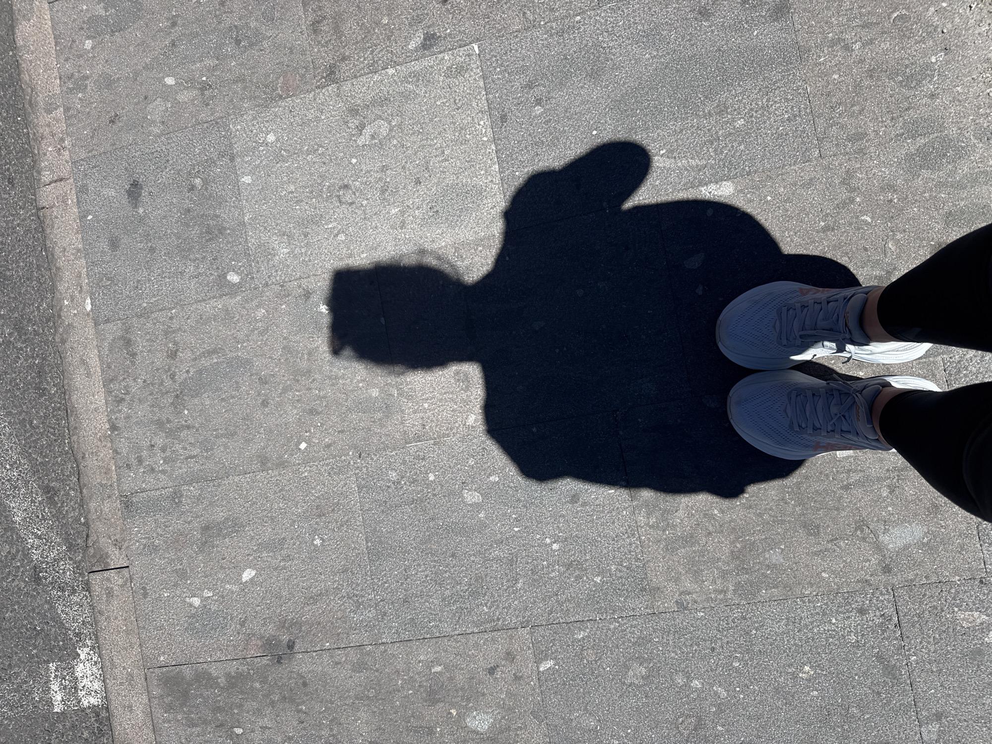 The image shows a pair of feet wearing light gray athletic shoes and black leggings, casting a shadow on a gray paved sidewalk. The shadow, originating from a person not fully visible, resembles a person with their arms raised, perhaps in a gesture of victory or greeting. The sidewalk is composed of large, rectangular stone tiles, and a portion of a darker asphalt surface with white markings is visible on the right side of the frame, suggesting the edge of a road or pathway. The environment appears to be an outdoor urban setting, possibly in La Orotava, Spain, given the sunlit conditions and typical street paving materials. The lighting indicates it is daytime, with a strong light source casting sharp shadows.