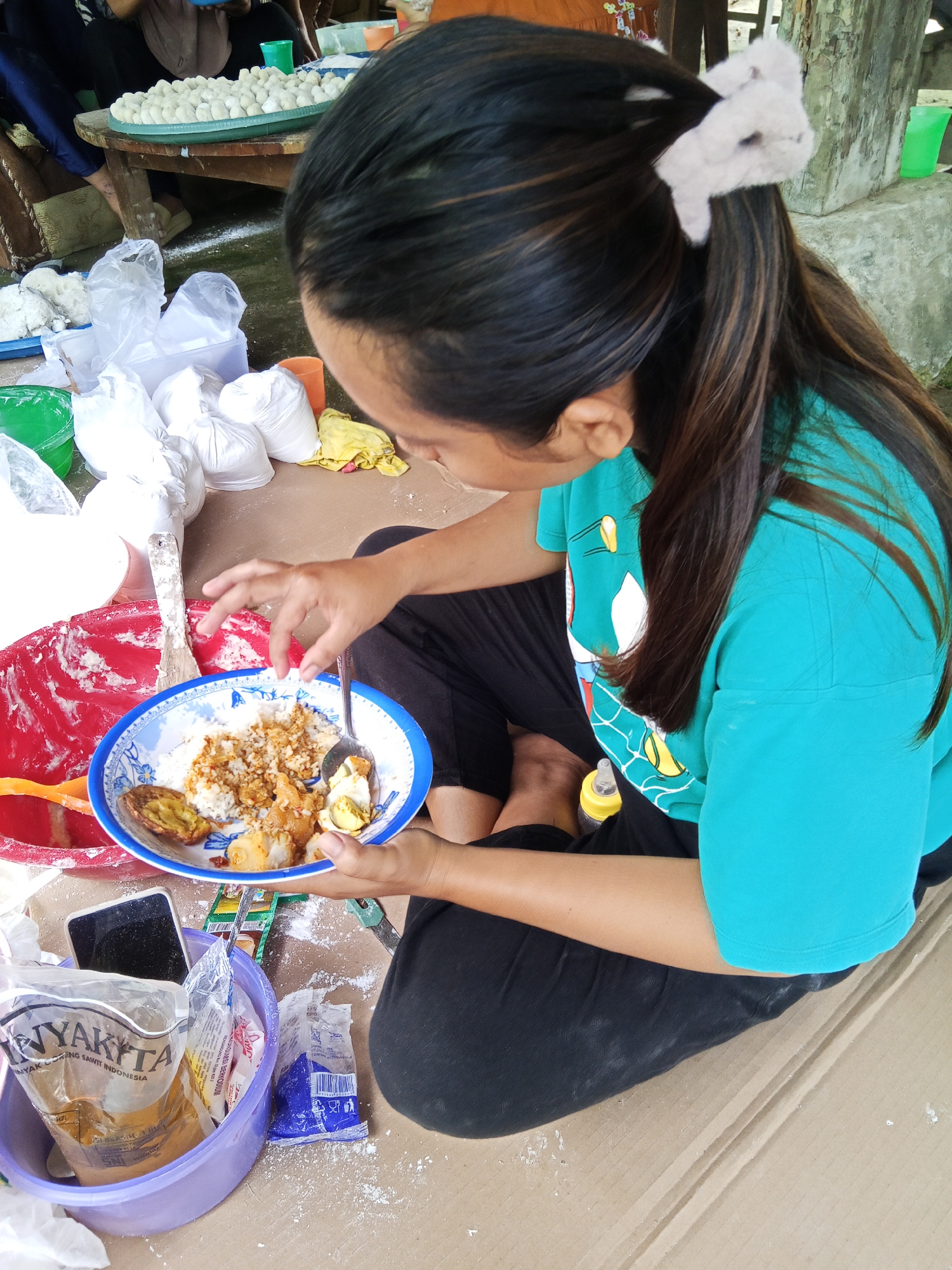A young woman in a turquoise t-shirt and black pants is sitting on the ground, eating from a blue and white bowl of rice with various food items. She is holding a spoon in her right hand and appears to be in the process of taking a bite. Next to her, to her left, is a large red bowl with white residue, a wooden spatula, and an orange plastic object. To her right, on the ground, is a purple plastic container filled with bags of what appears to be cooking ingredients, a smartphone, and a small yellow bottle. The ground is covered in white powder, likely flour or a similar ingredient, suggesting food preparation is taking place. In the background, a table is laden with a large tray of small, round white food items, and several plastic bags filled with white powder are scattered around. The setting appears to be outdoors, possibly a yard or a rural area, given the natural light and rough texture of the ground. The overall scene suggests a communal food preparation or cooking activity.