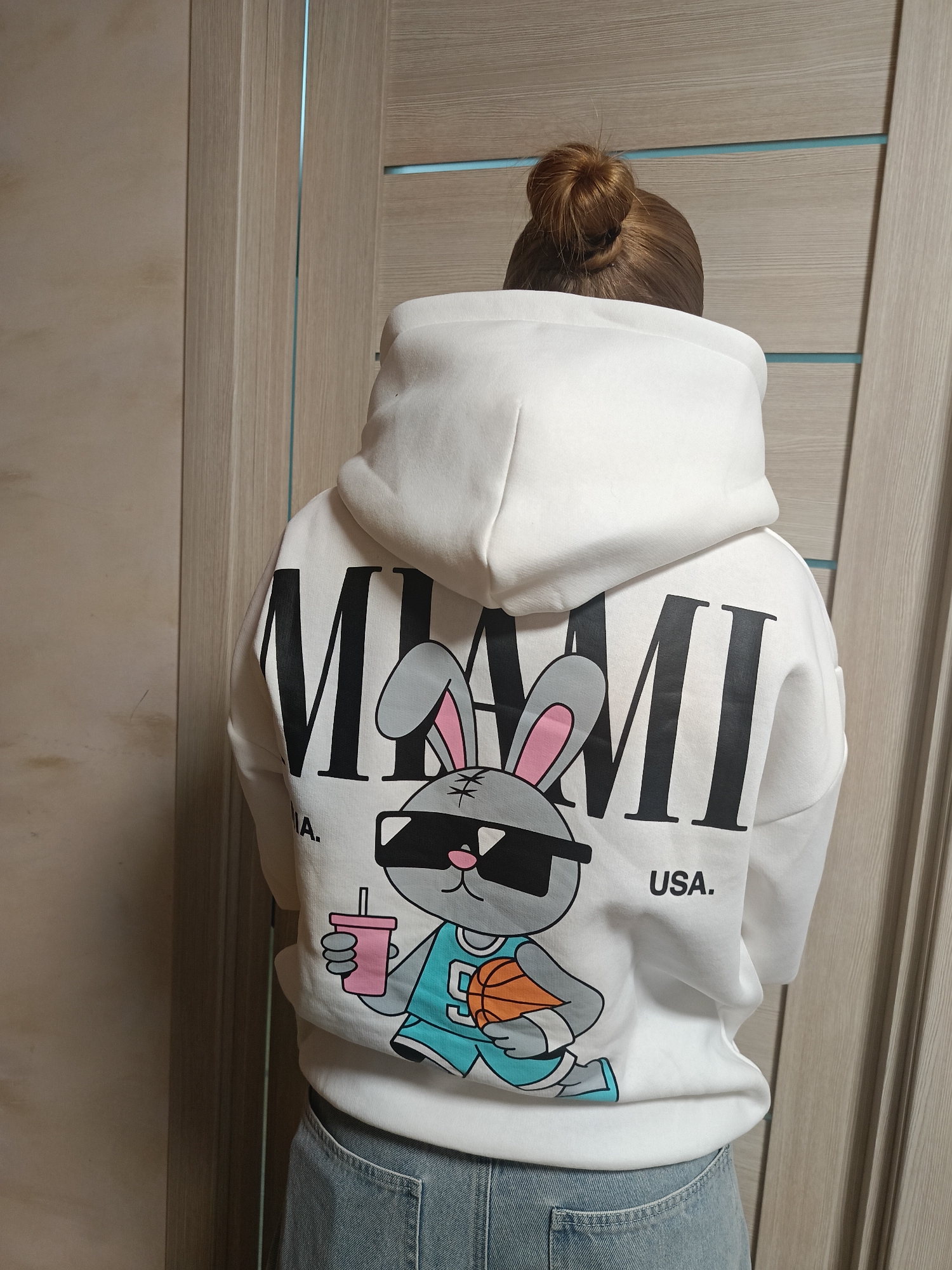 A person is standing indoors, wearing a white hoodie with a cartoon bunny printed on the back. The bunny is wearing sunglasses and a basketball jersey, holding a drink in one hand and a basketball in the other. The word "MIAMI" is printed above the bunny, and "USA." is below it. The person has their hair in a bun. The background is a light-colored wooden door. The location is unknown, not necessarily Chaikovsky, Russia.