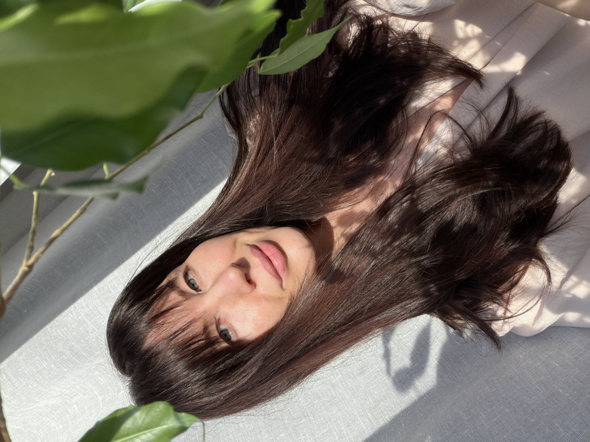 A woman with fair skin and long, dark brown hair lies on a light gray textured surface. Her face is framed by her hair and she gazes towards the camera with light-colored eyes. A green plant is visible in the top left corner of the frame. The scene is illuminated by bright natural light, creating distinct shadows. The photo appears to have been taken at an angle. There is no verifiable information regarding the location of this photo to be Perm, Russia.
