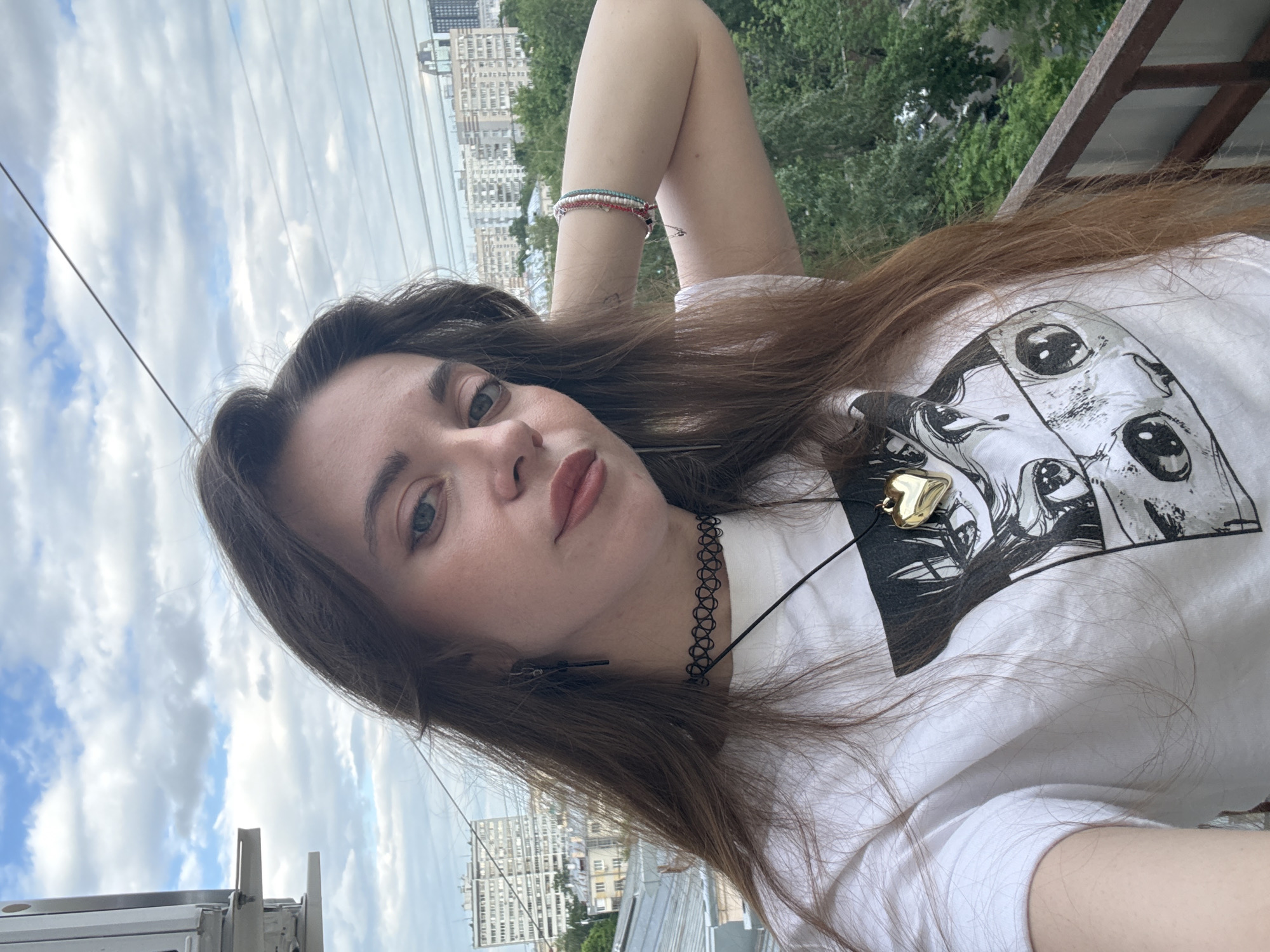 A young woman is lying down on a balcony or rooftop in Moscow, Russia. She is facing the camera. Her head is tilted slightly. She is wearing a white t-shirt with a black-and-white manga print. A gold heart-shaped pendant necklace is visible. The woman has brown hair and is wearing a black choker. Her right arm is bent behind her head and she wears a multiple bracelets. In the background are apartment buildings and trees typical of the Moscow urban landscape. The sky is visible with scattered clouds.