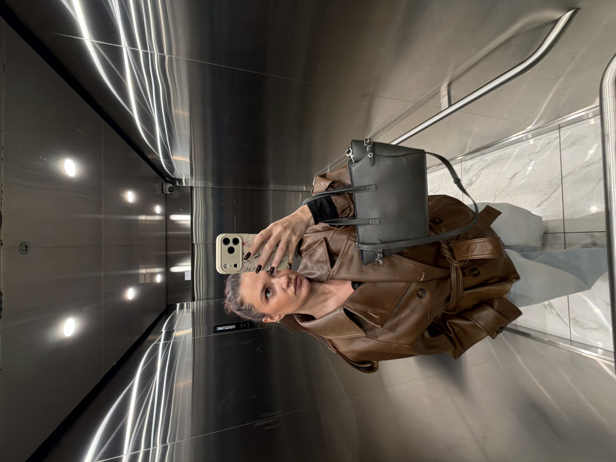 A person is taking a selfie in what appears to be an elevator. The person is wearing a brown leather jacket and holding a cell phone in their right hand with a gray handbag hanging from their shoulder. The elevator has reflective walls and a marble tile floor. There are lights on the ceiling. The location is unspecified, but the image suggests an urban setting. There is no evidence in the photo to suggest the city of Belgrade, Serbia.