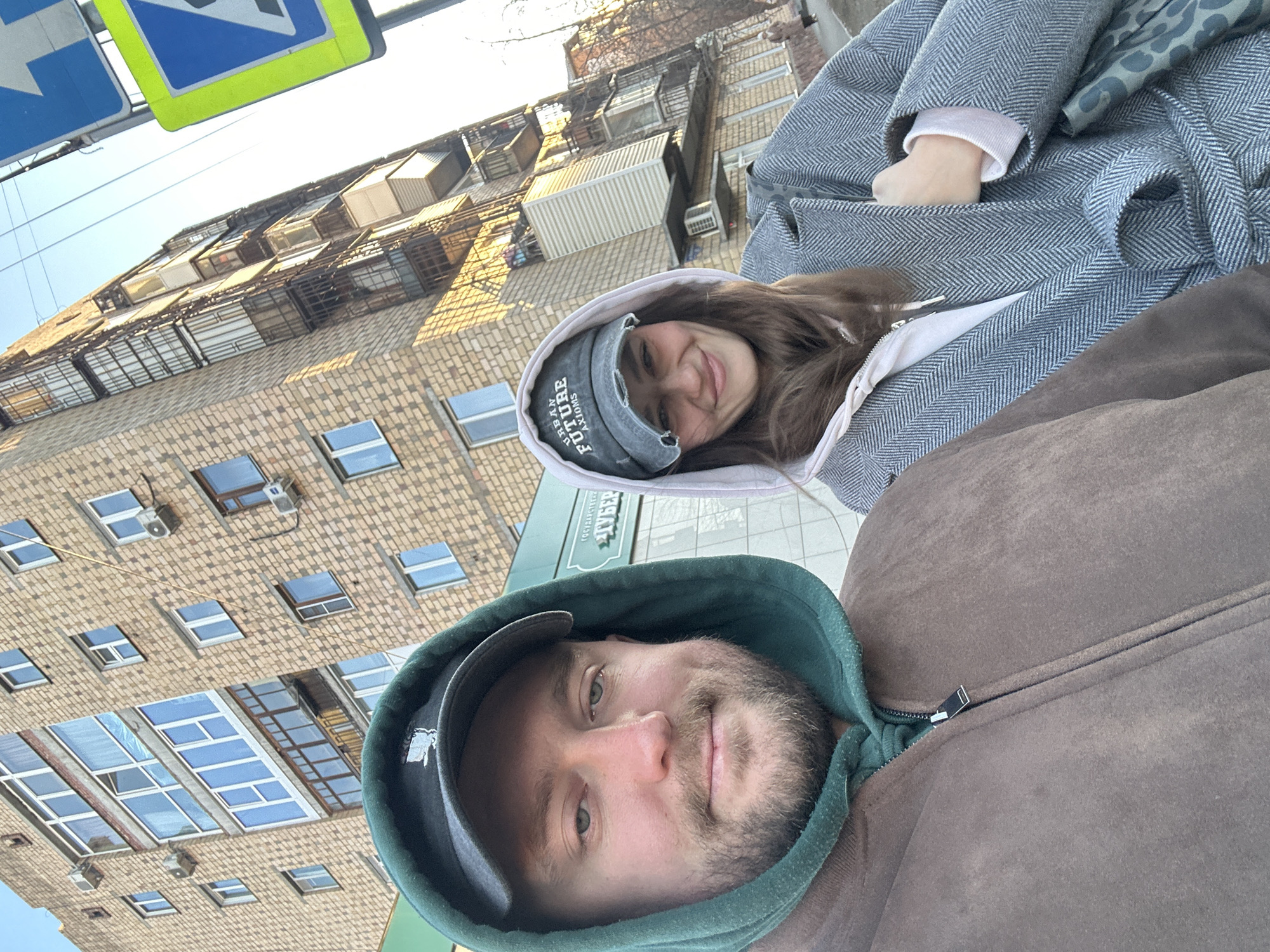 A selfie taken by a light-skinned male wearing a zipped brown hoodie with a green hood and a black baseball cap is visible. The image also shows a light-skinned female, smiling and wearing a white hoodie, a grey baseball cap, and a grey patterned coat. The background includes a multi-story building with numerous windows, and a street sign in the upper left. Based on the Cyrillic characters on a sign in the background, the location is likely Krasnoyarsk, Russia.