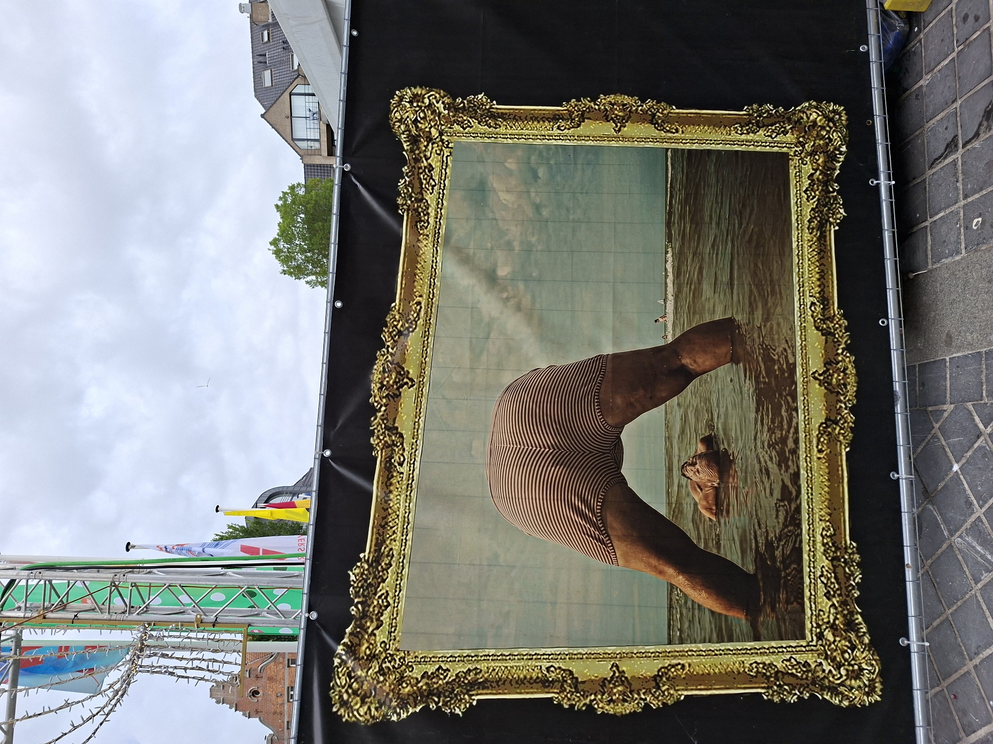 The image shows a large banner with a framed artwork depicting a person with their lower body submerged in water, head down, with only their striped shorts visible above the water. The artwork is framed by an ornate, golden frame. In the background of the artwork, there is a cloudy sky and a calm sea with a few distant figures and sailboats. Above the banner, the cityscape of Ostend, Belgium, is visible, including buildings and flags. A large, green, metal structure with flags and string lights is also present, suggesting a festival or event. The banner is attached to a metal railing, and the foreground shows a cobblestone pavement.