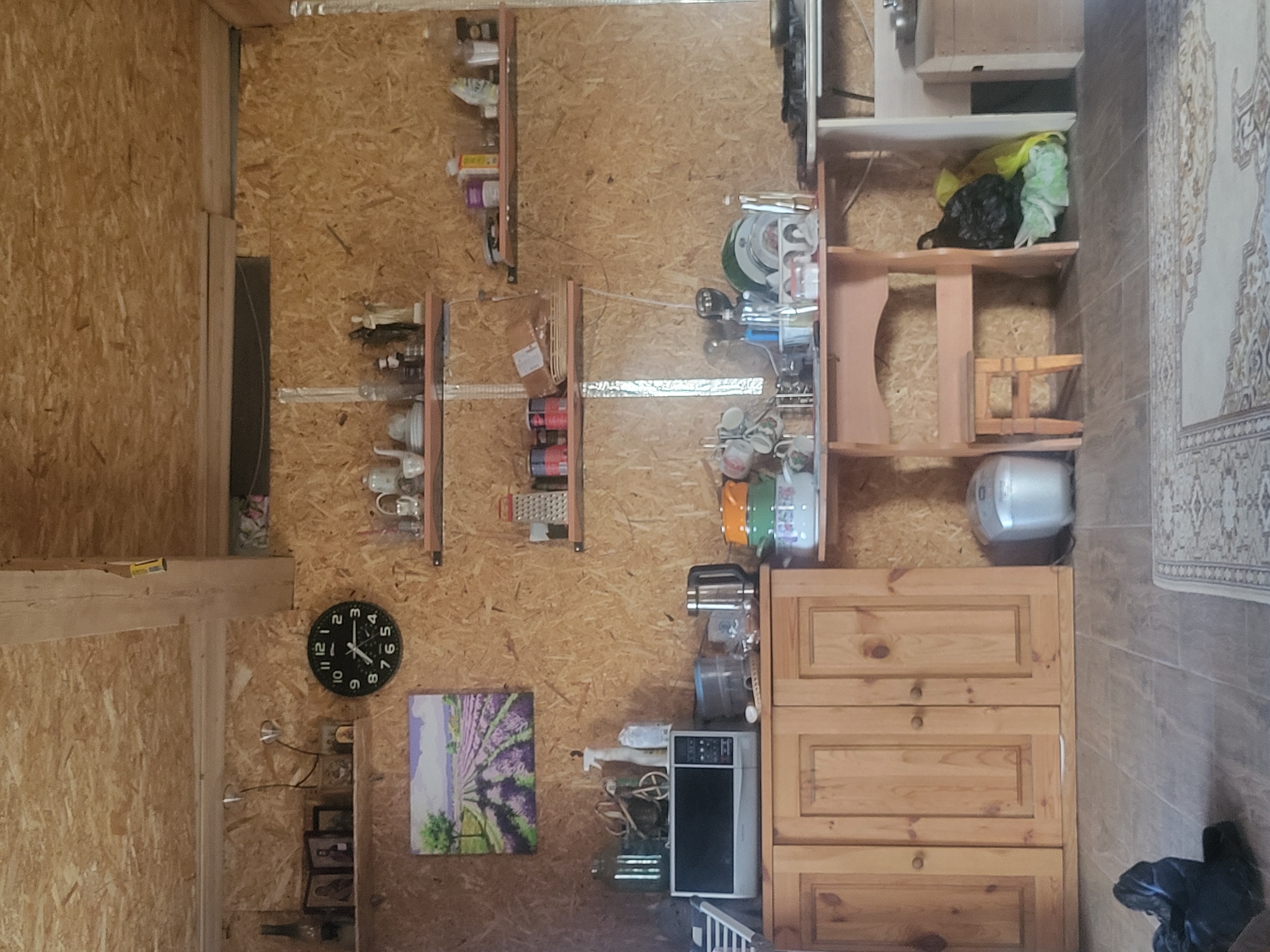 The image depicts the interior of a rustic kitchen area. The walls and ceiling are constructed from wood particle board, with exposed wooden beams. On the wall, a circular clock with black Roman numerals is visible. To the left of the clock, a framed painting of a lavender field and trees hangs above a microwave. Shelves are mounted on the wall, displaying various kitchenware and decorative items, including pitchers, bowls, and small figurines.

A wooden cabinet with two sets of double doors occupies the left side of the lower portion of the image. Above the cabinet, a microwave sits on top, next to a spray bottle and other kitchen items. To the right of the cabinet, a wooden table with a stool is positioned, with dishes and kitchen utensils on it. Below the table, a silver rice cooker is placed on the floor.

The floor is tiled, and a patterned rug is partially visible in the foreground. In the lower right corner of the image, a black trash bag and some green and yellow plastic bags are visible. The overall impression is that of a functional, somewhat cluttered, domestic space with a focus on practical storage and display. The specific location in Сцяпанкаўскі сельскі Савет, Belarus is not visually evident from the image itself.