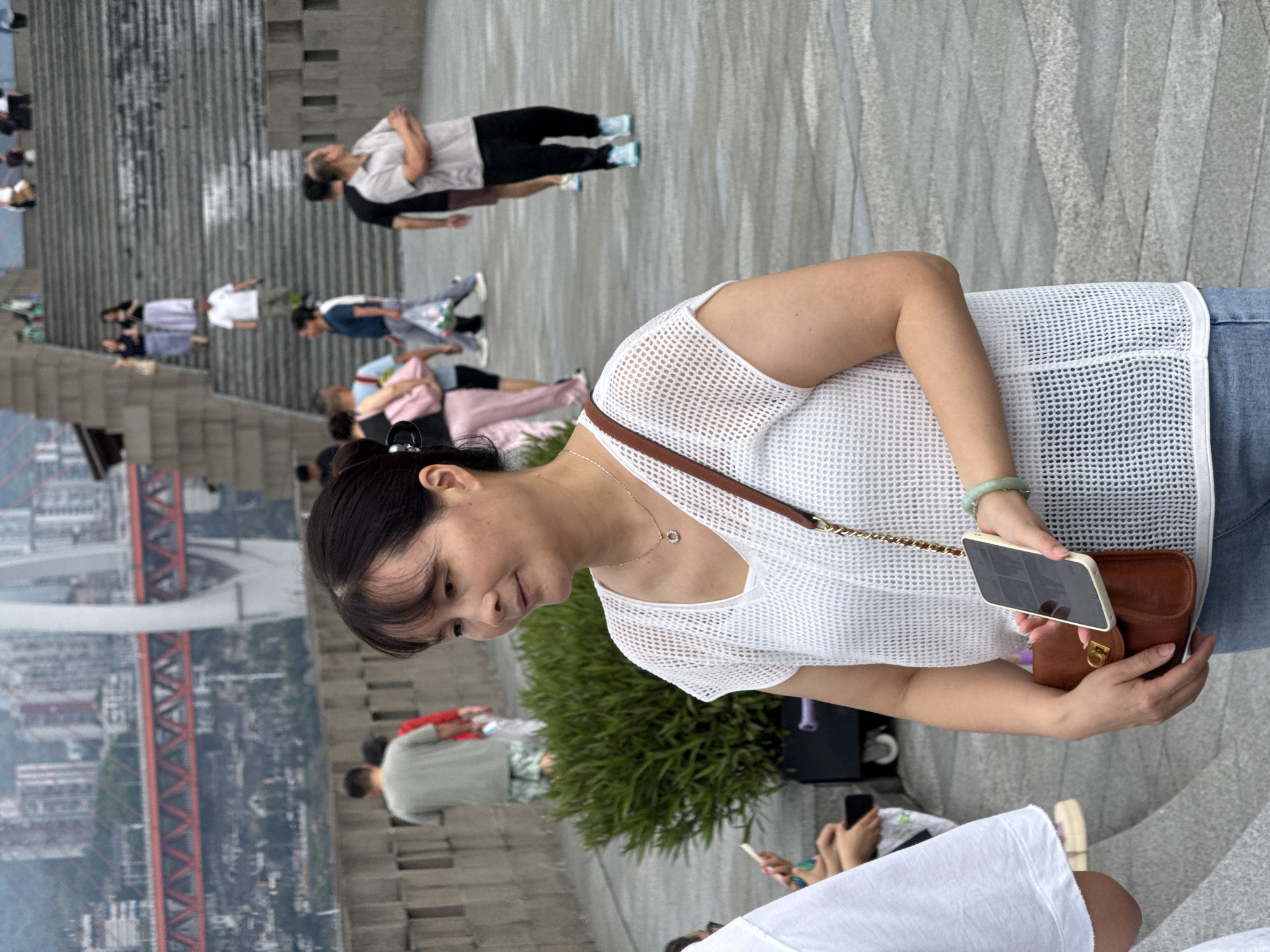 In Nan'an District, China, a woman with dark hair and a white, open-knit top stands in the foreground, holding a smartphone and a small brown purse with a gold clasp. She is wearing a jade bracelet on her right wrist. Behind her, a paved walkway and a large bush are visible. In the background, people are ascending a wide stone staircase leading to a city skyline with modern buildings and a red suspension bridge. The overall scene captures a moment in an urban public space.