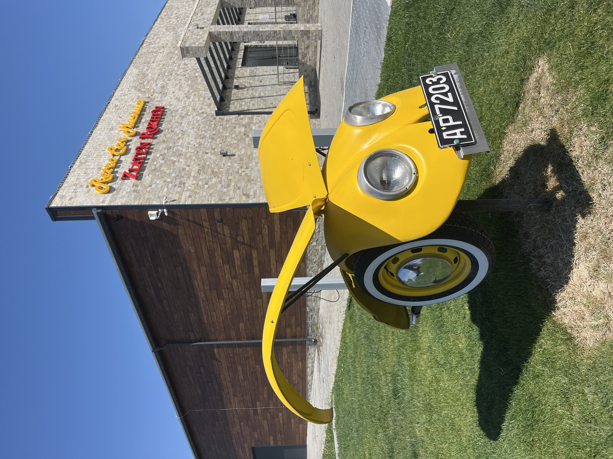 The image depicts a partially visible, bright yellow car, possibly a Volkswagen Beetle, with the license plate "AP7203", parked on a green lawn. The car's hood is open. A building with a facade of light-colored stone and dark wooden panels is visible in the background. The building's name, "Zlaten Rozhen Museum", is displayed in yellow and red letters. The image was captured during daylight under a clear blue sky. The location is Kapatovo, Bulgaria, indicated by the name on the building. The shadow of a person taking the photo is visible on the grass.