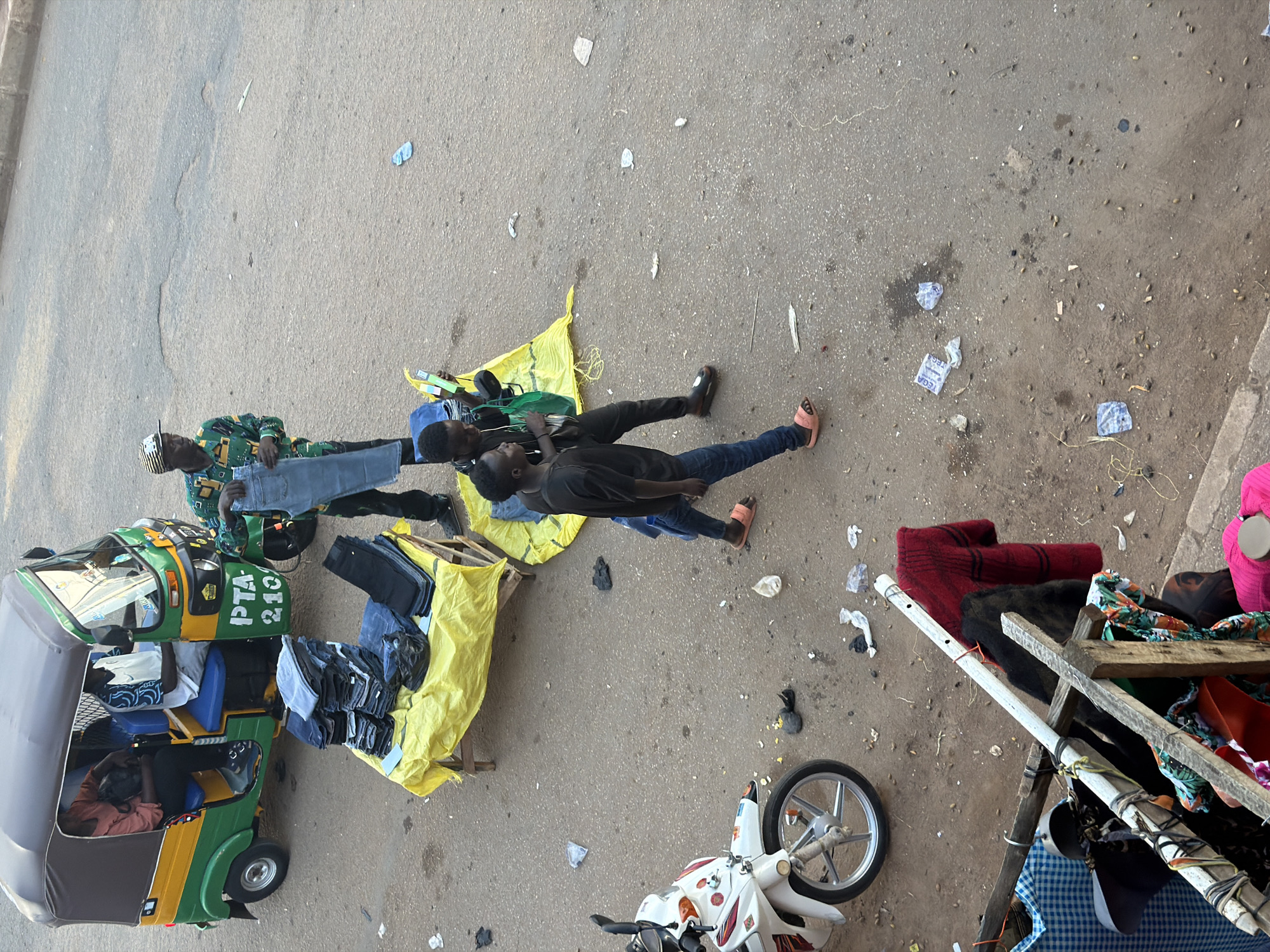 In Bukuru, Nigeria, a street scene unfolds with a focus on commerce and daily life. In the foreground, a motorcycle is parked, its white body a contrast to the dusty, paved ground littered with debris and small objects. To the right, a makeshift stall displays various garments, including a red sweater and patterned fabrics, suspended from wooden and metal poles.
Further into the scene, a bright yellow tarp covers a wooden table laden with stacks of folded denim jeans. Two young men stand near the table. One, in a black t-shirt and jeans, holds a mobile phone, and the other, similarly dressed, is examining a pair of jeans. In the background, a colorful auto-rickshaw, painted green, yellow, and red, is parked. A man in a patterned shirt stands beside the rickshaw, holding up a pair of jeans. Two individuals are seated inside the rickshaw, one wearing a reddish-orange shirt and the other a blue and white patterned shirt. The ground is a mixture of asphalt and dirt, with more debris scattered throughout. The lighting suggests it is daytime, possibly late afternoon, given the shadows.