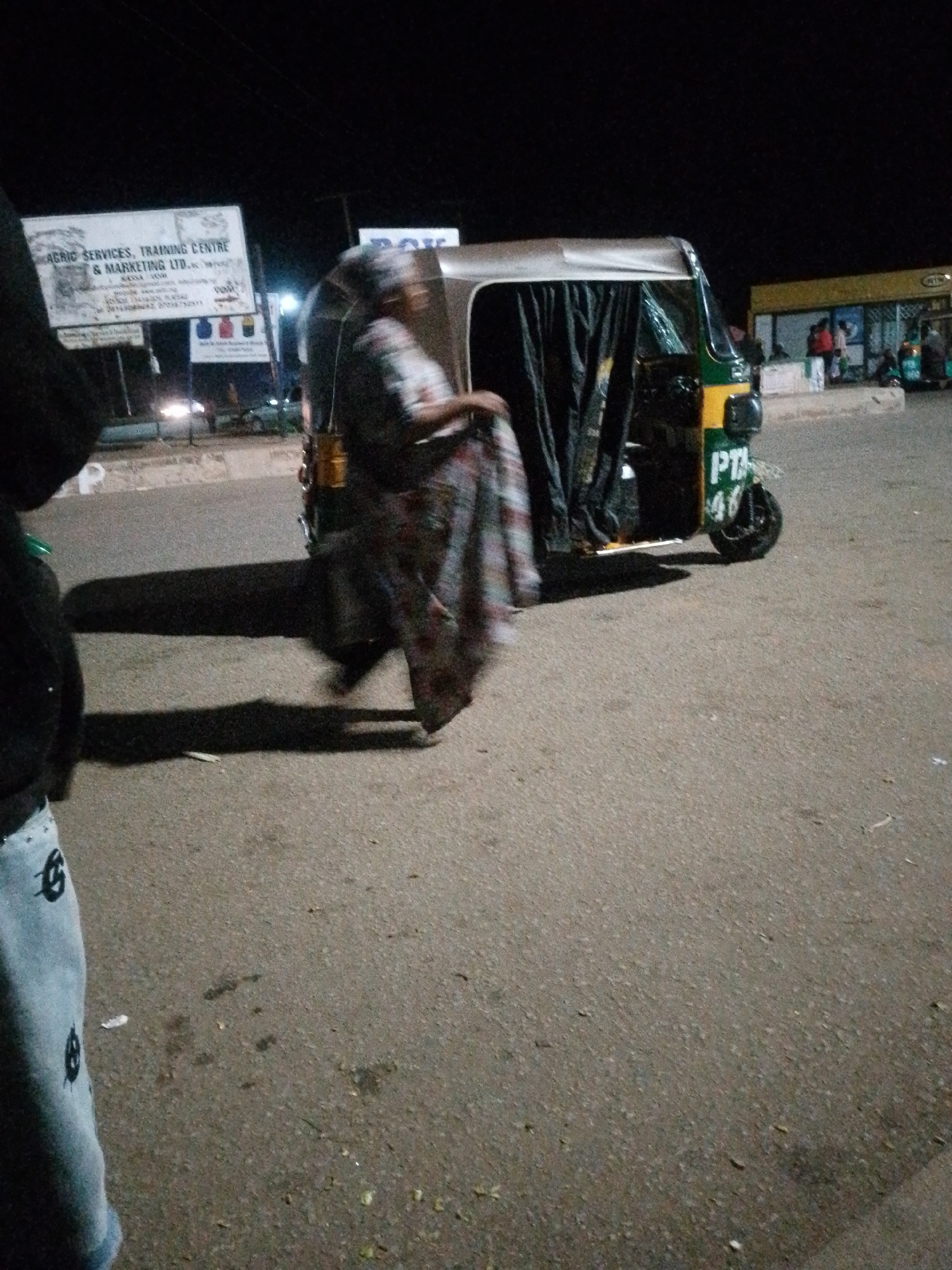 At night in Bukuru, Nigeria, a woman in a patterned wrap is standing next to a green and yellow auto-rickshaw. The auto-rickshaw has a license plate that reads "PTA 46". In the background, a sign advertises "AGRIC SERVICES, TRAINING CENTRE & MARKETING LTD". Other people are visible in the distance near a building with a yellow awning. The ground is asphalt.