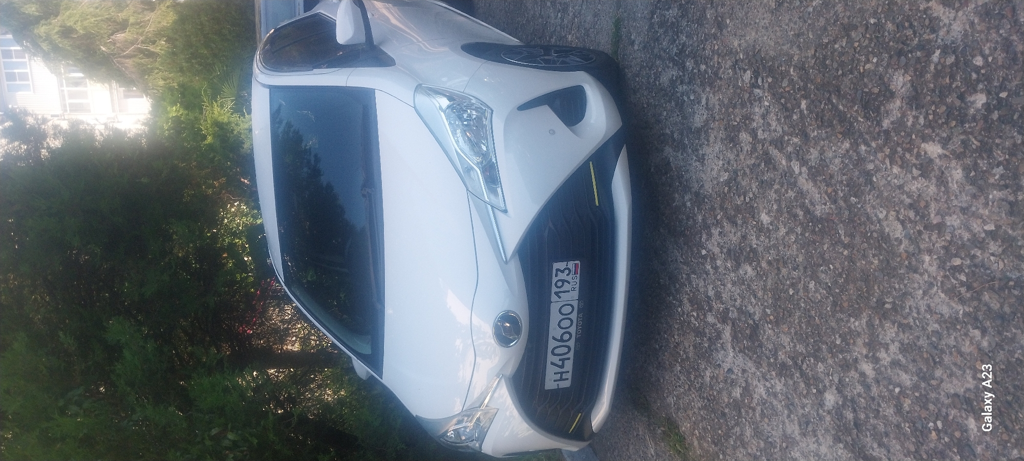 A white Toyota Vitz with the license plate H40600 193 is parked on a gravel surface in front of dense green trees and a light-colored building. The car's front end is visible, showing the headlights, grille with the Toyota emblem, and the license plate. The scene appears to be a street in Tuapse, Russia, given the presence of Russian license plates and the characteristic architecture of the building. The lighting suggests it is daytime.