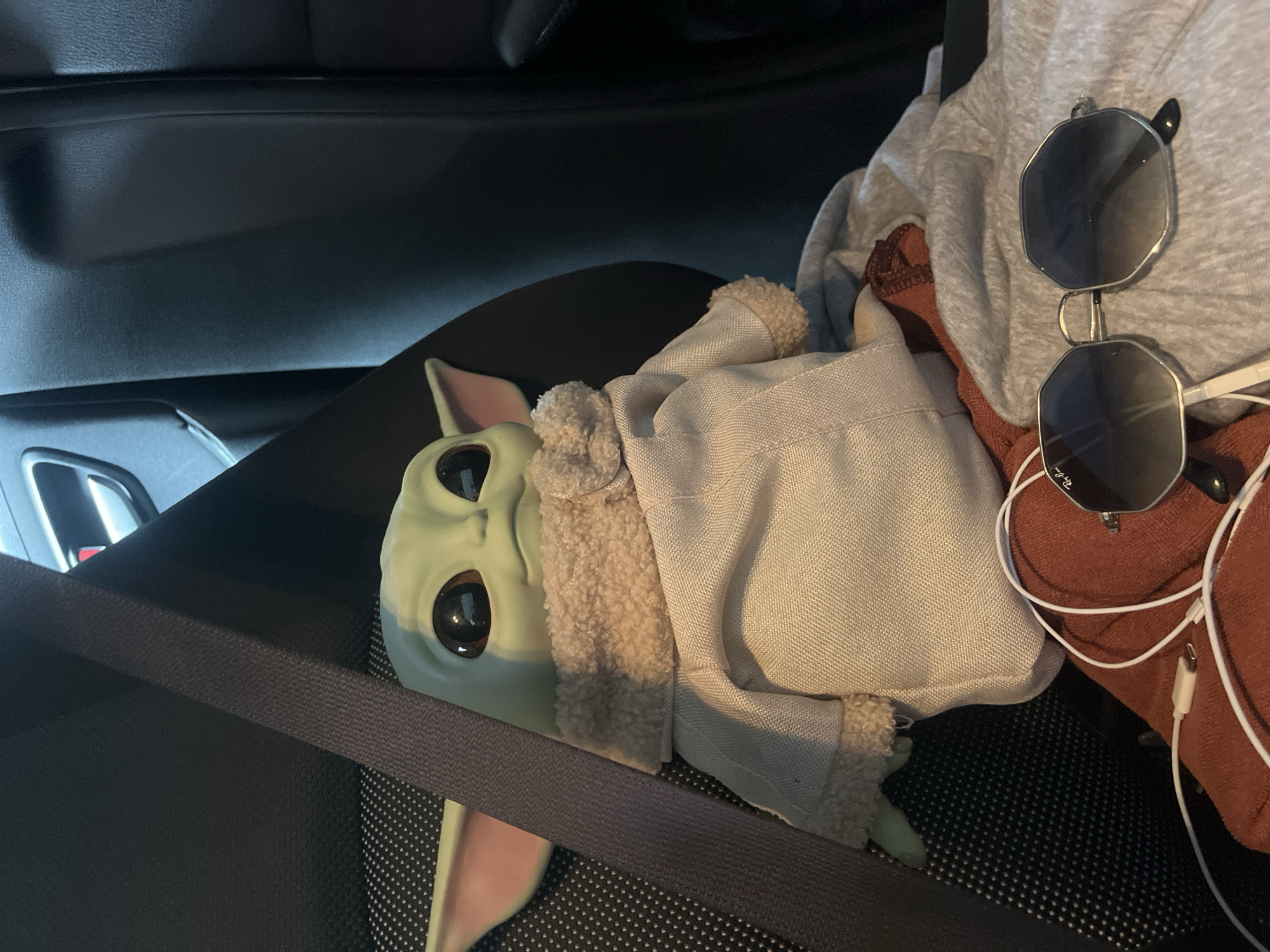 In Rawai, Thailand, a plush toy resembling the character Grogu from Star Wars is seated in the passenger seat of a vehicle. The toy is secured by a dark gray seatbelt. Adjacent to the toy, on the seat, are a pair of octagonal, silver-rimmed sunglasses with gradient lenses, resting on a beige sweatshirt and a rust-colored cloth. White charging cables are also visible on the seat, one connected to a white adapter. The background consists of the car's dark interior, including a black seat and a portion of the door panel.