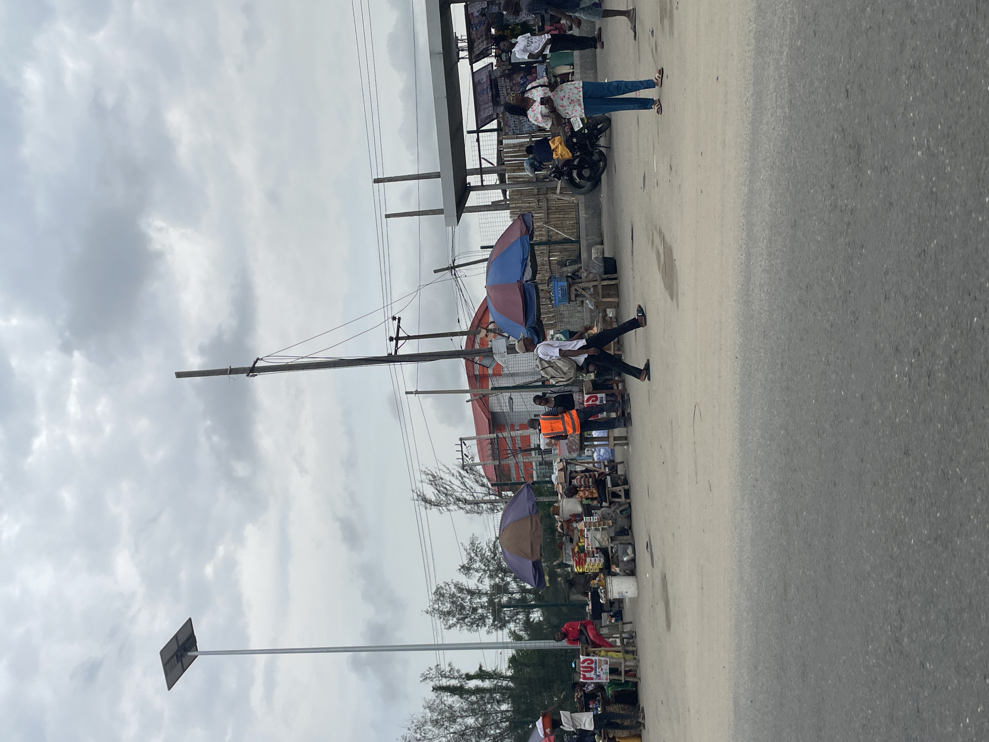 The image depicts a street scene in Lagos, Nigeria, on an overcast day. The street is lined with informal market stalls covered by umbrellas and makeshift shelters. Vendors and pedestrians are present, and a few are interacting with the stalls. One person is crossing the street. Overhead electrical lines and utility poles are visible. A bus stop structure is also present. The road is paved, and the sky is cloudy.