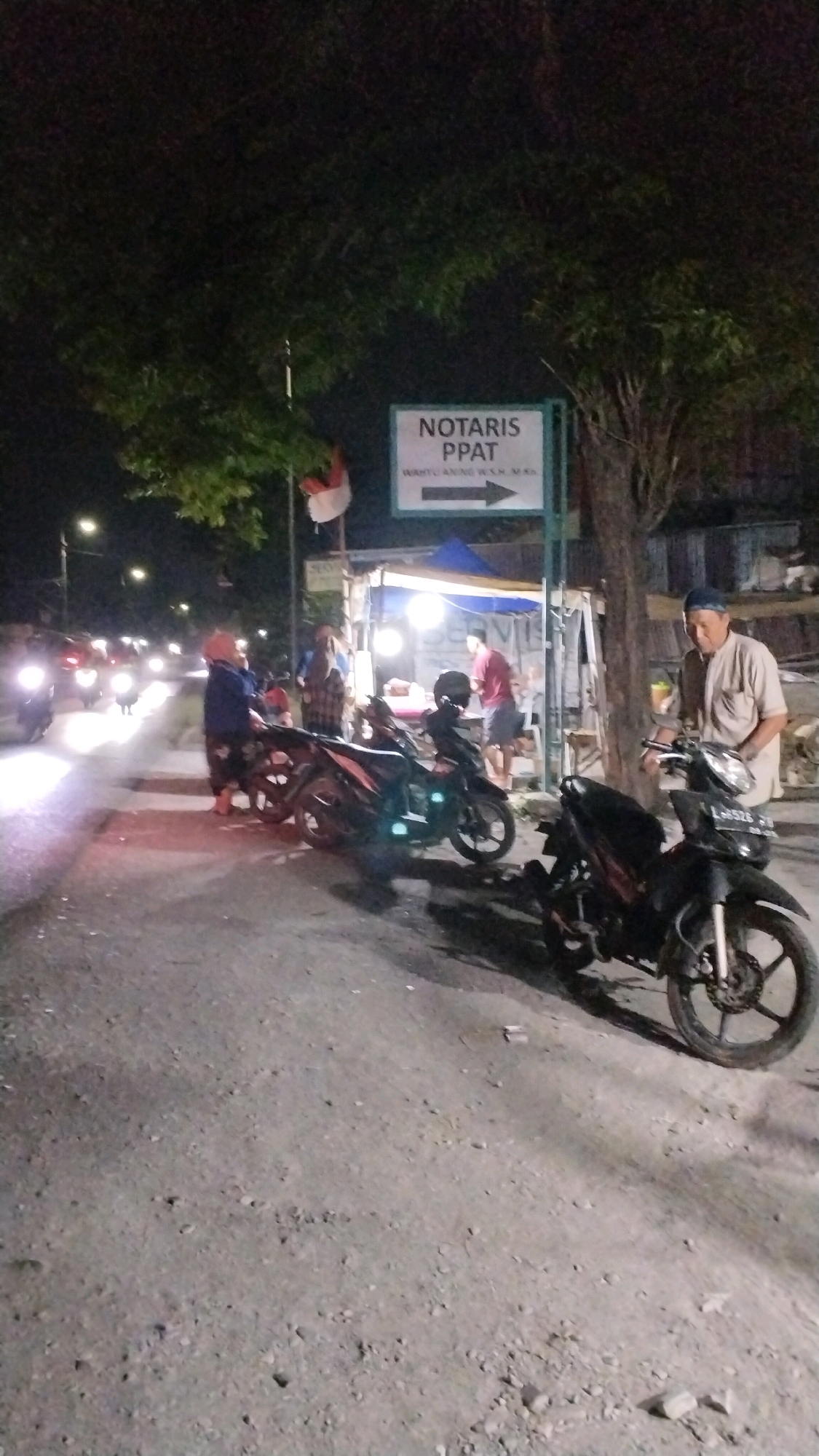 A nighttime street scene in Canggu, Indonesia, shows a busy road with several motorcycles parked and moving. A sign with "NOTARIS PPAT" is visible, indicating a notary public's office. People are gathered near a stall with bright lights, possibly a food or service vendor. The ground is a mix of asphalt and gravel. Trees overhang the scene, and the sky is dark.