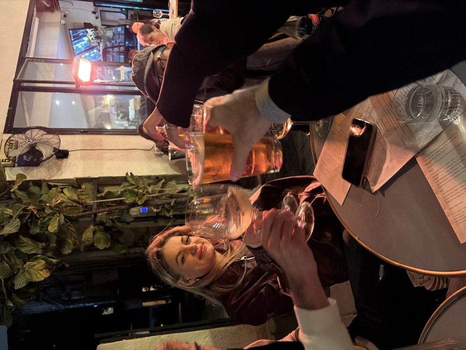 The image depicts a nighttime social gathering at an outdoor or semi-outdoor bar or restaurant in Tel-Aviv, Israel. This location is confirmed by Hebrew text on a visible Coca-Cola bottle and a partial street sign with the number "5" and Hebrew script on a wall.
At least five individuals are present, participating in a toast. A blonde female, centrally positioned, wearing a dark reddish-brown leather jacket, smiles towards the camera while holding a glass of white wine. Her right hand is raised, clinking glasses with others. The hands and arms of at least four other individuals are visible, holding glasses containing beer, white wine, and other beverages, indicating a collective toast. One participant wears a dark flat cap, visible on the right side of the frame. Another woman is partially visible in the background, seated at a separate table.
The foreground features a round, light-colored table covered with paper menus or receipts. A black smartphone with a metallic frame lies face down on the table. An empty clear drinking glass is also present.
The background shows a light-colored wall adorned with lush green, large-leafed plants. A wall-mounted oscillating fan is affixed to this wall. An overhead red radiant heater is mounted near a window or doorway, reflecting light. Through the window, an interior space with a television screen displaying blue and white patterns is visible. A Coca-Cola bottle with Hebrew text is visible on a table in the background. The lighting is artificial and generally warm, creating a convivial atmosphere.