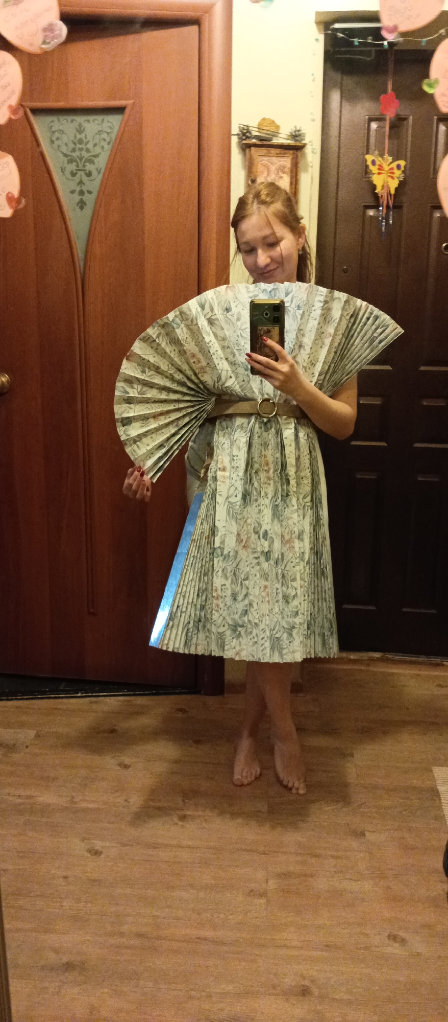 A person is standing on a wooden floor in front of two wooden doors. The person is holding a smartphone in their right hand, presumably taking a photo of themselves. They are wearing a pleated dress or skirt with a floral pattern and a tan belt with a large buckle. A fan-like object made of the same material as the dress is wrapped around the upper body. The person is barefoot. The background includes wall decorations on the door to the right and a mirror reflection is visible, suggesting the setting might be indoors, possibly a home. The location appears to be Unknown, Unknown.