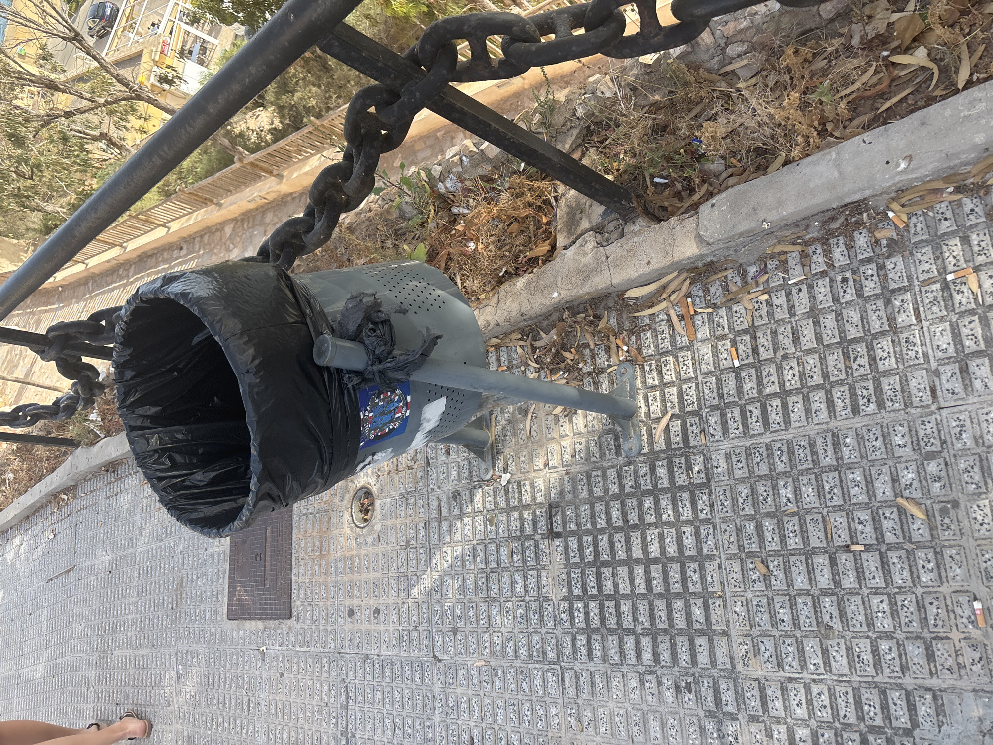 A high-angle, close-up shot shows a metal trash can on a textured concrete walkway. The trash can is mostly empty and lined with a black plastic bag. A thick metal chain is attached to the trash can and runs along a railing. The walkway is littered with cigarette butts and dried leaves. In the background, a metal railing runs alongside a path with a wooden fence. Beyond the path, trees and buildings are visible, suggesting an outdoor urban environment. A person's legs and feet are visible on the left side of the frame, wearing sandals.
