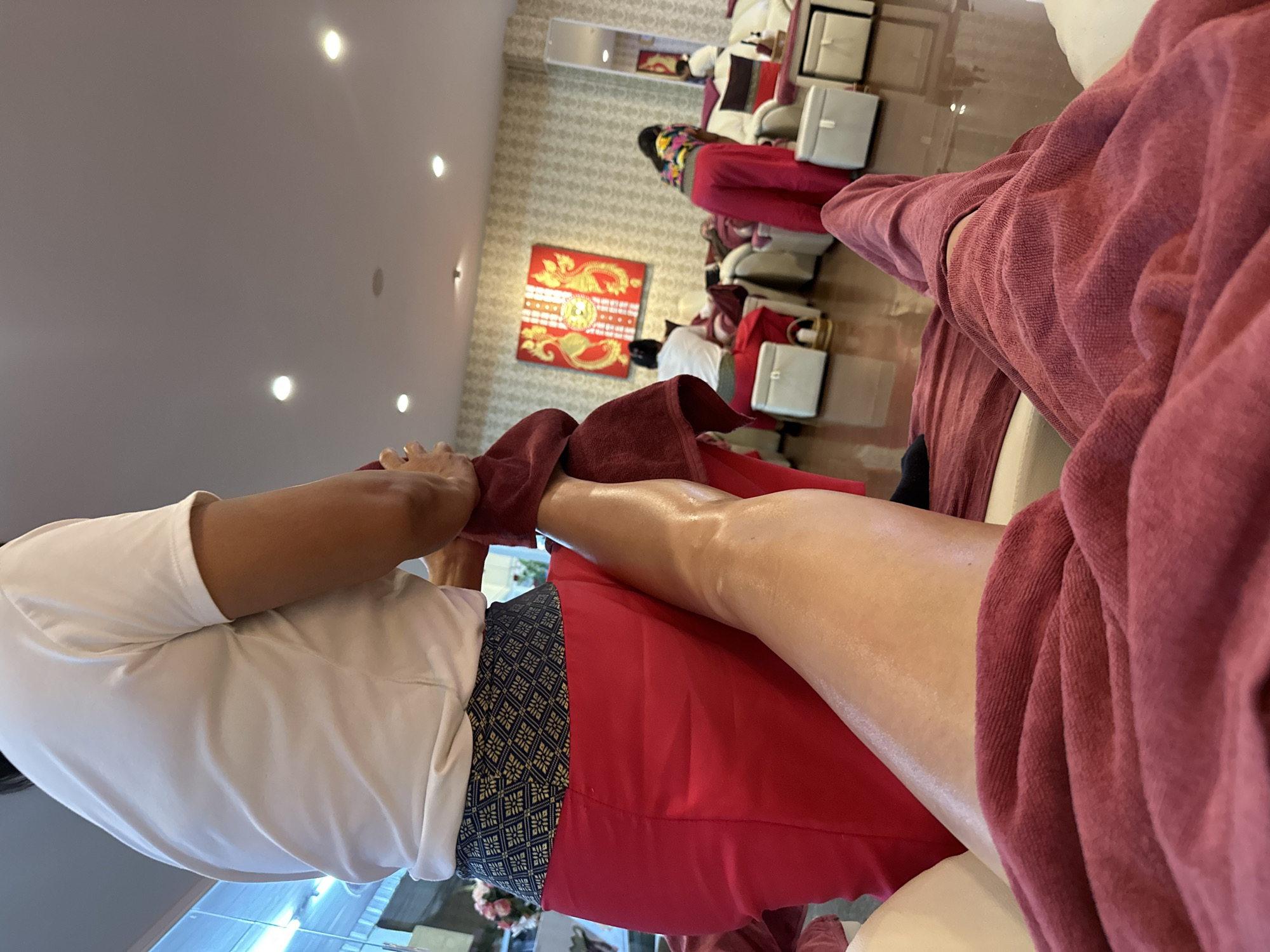 The image shows a person receiving a foot massage, potentially in Pattaya City, Thailand, given the prevalence of such services in the area. The person receiving the massage is wearing loose, reddish-pink clothing and is seated in a cushioned chair. A masseuse, dressed in a white shirt and a traditional Thai-style red sarong with a geometric pattern around the waist, is working on the person's leg. The leg appears to be oiled and is draped with a reddish-brown towel.
In the background, there are other patrons receiving similar treatments in a room adorned with Thai-style decorations, including a large, framed piece with golden dragon motifs on a red background. The room is illuminated by recessed ceiling lights and features wallpaper with a subtle pattern. White pedicure stations are visible, along with the chairs people are sitting in. The overall setting suggests a professional massage or spa environment.