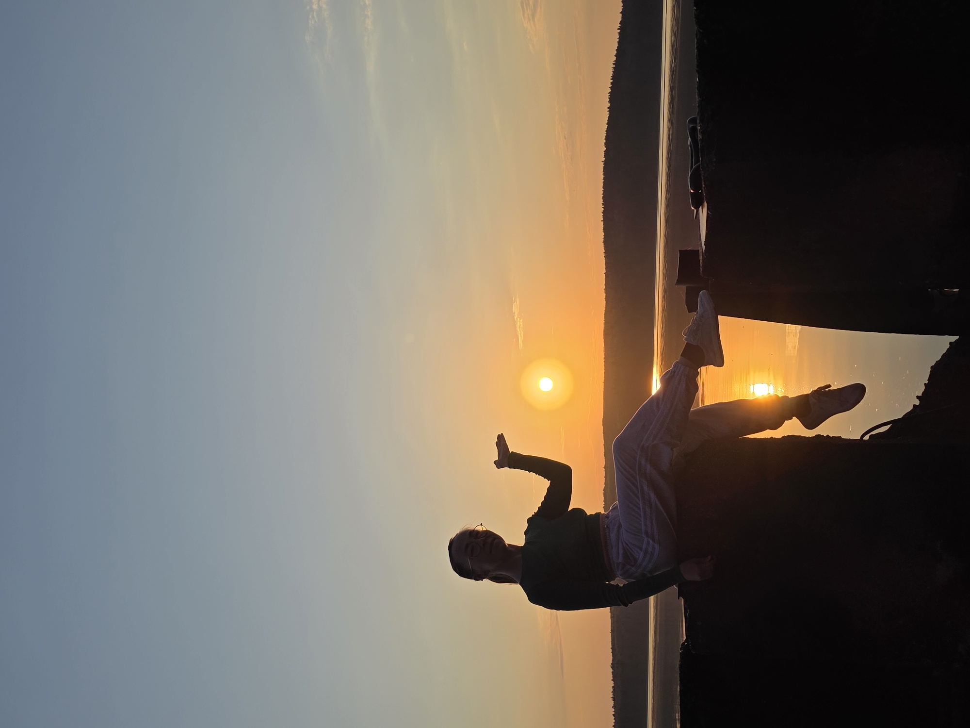 A silhouette of a woman is positioned to the right, against a backdrop of a bright orange sunset. She is sitting on a dark, solid surface, possibly a ledge or structure, with her legs extended towards the sunset. Her right arm is raised with her hand open. She is wearing glasses, long sleeves and pants. The sunset is over a body of water, with a dark, treeline visible in the distance, suggesting a lake or reservoir. The left part of the scene shows the sky, ranging from blue at the top to orange closer to the horizon. No information is provided to determine the image was taken in the city of Chaikovsky, Russia.