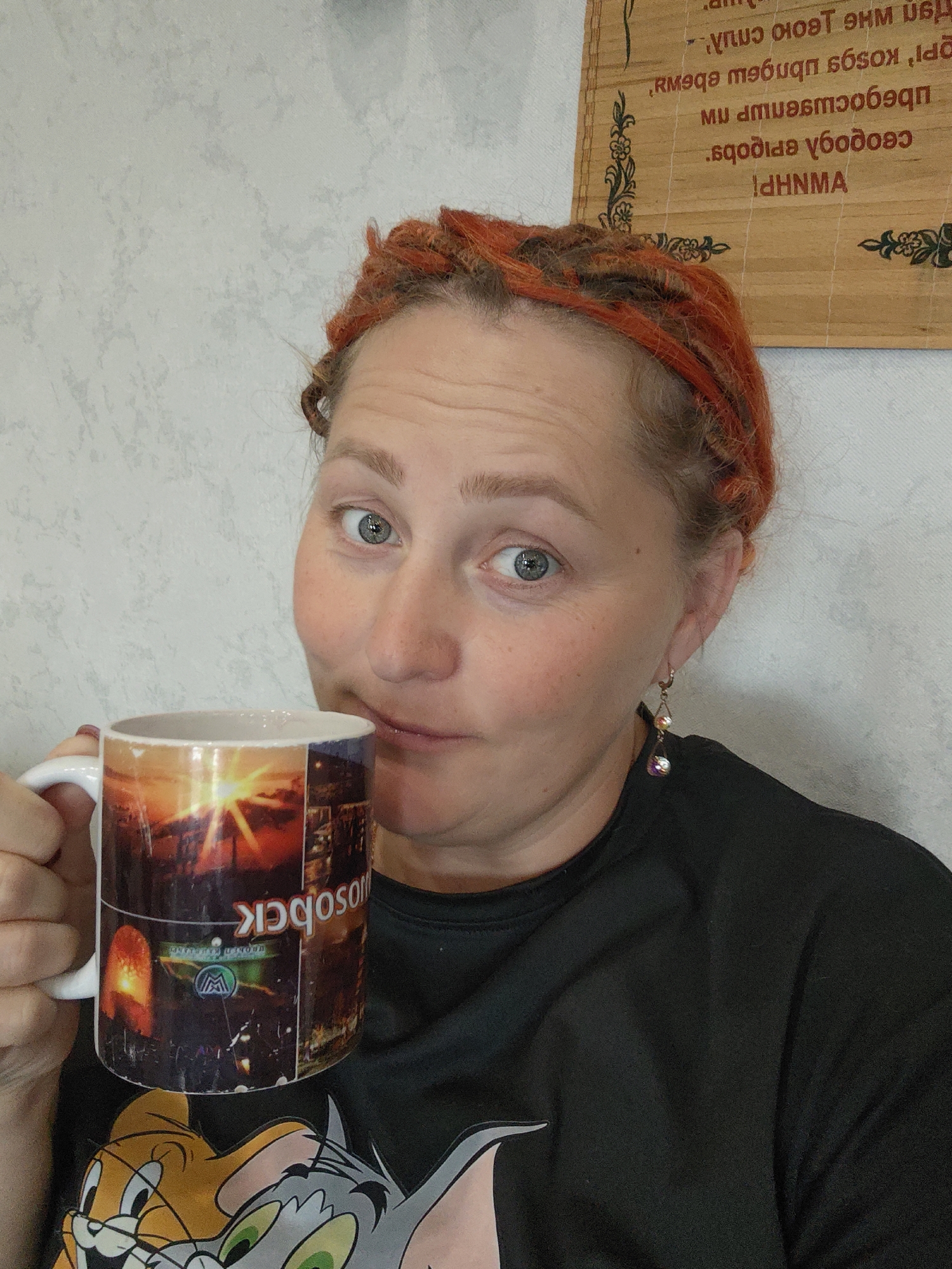The image shows a woman with reddish-orange dreadlocks holding a mug. The mug has an image of a city skyline at sunset and the word "Moscow" written on it. The woman is wearing a black t-shirt with a graphic of Tom and Jerry. In the background, a wooden plaque with some text in Cyrillic script is visible. The wall behind her appears to be textured and light gray. The text on the wooden plaque is in a mirror image and appears to be some sort of inscription or proverb. The lighting in the image is natural, coming from the front and slightly to the left. There are no indications of the location being in the city of Mokino, Russia.