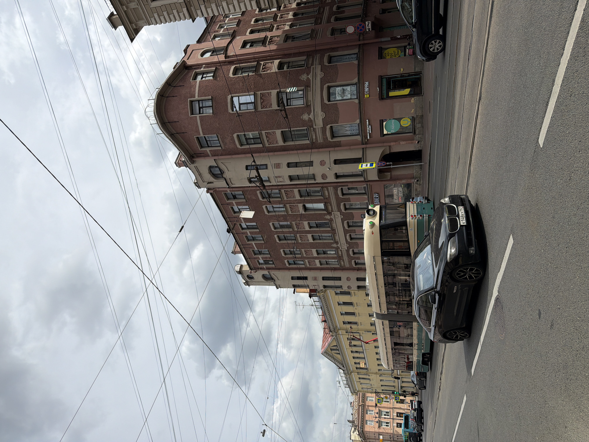The image depicts a street scene in Saint Petersburg, Russia, characterized by overcast skies and a collection of urban buildings. A black sedan is in the foreground, driving on an asphalt road with white lane markings. Behind it, a tram with a light-colored exterior and green accents travels on tram tracks. Several multi-story buildings with intricate architectural details line the street. The buildings are a mix of colors, including pink, yellow, and brick red, with many windows. Overhead, a dense network of power lines crisscrosses the sky. The overall impression is a bustling city street on a cloudy day.