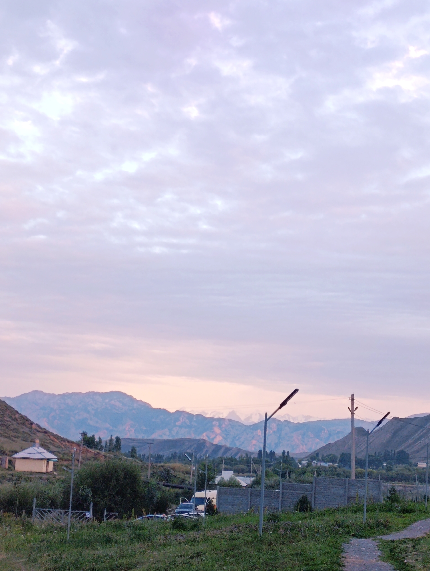The image depicts a rural landscape in Kazan-Kuygan, Kyrgyzstan, under a pale, cloudy sky with hints of pink and purple from the twilight. In the foreground, there is a path made of gravel, bordered by green grass and foliage. Several lampposts are visible, some with active lights. A grey concrete fence runs horizontally across the mid-ground, with a white minivan and a dark car parked behind it. Further back, a small, yellow, house-like structure with a white roof is nestled on a hillside. Beyond that, a range of mountains dominates the background, their slopes a mix of muted greens, browns, and blues, with some snow-capped peaks visible. The overall impression is one of serene natural beauty in a quiet, undeveloped area.