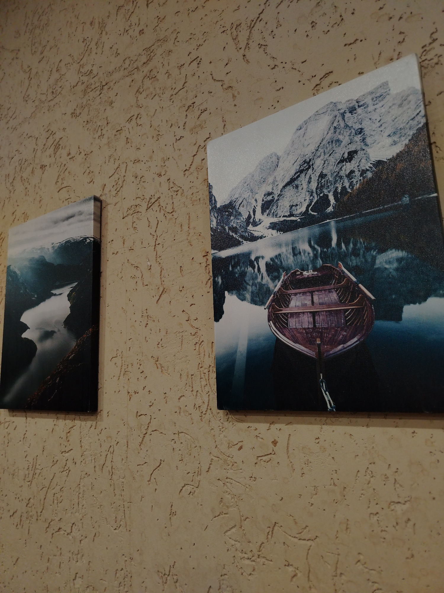 The image captures an indoor scene in Chaikovsky, Russia, featuring a light beige or sandy-colored wall with a distinctive textured finish, possibly stucco or a similar decorative plaster, characterized by subtle, irregular raised patterns. Two canvas-mounted artworks are displayed on this wall.
On the right, a larger rectangular canvas presents a serene natural landscape. The foreground features a traditional wooden rowboat, tethered and resting on calm, dark water. The water perfectly reflects snow-capped mountains under an overcast sky, creating a striking mirror image. The mountains are rugged and substantial, with patches of snow contrasting against darker rock and sparse vegetation. The color palette is dominated by cool blues, grays, and whites for the landscape, with the boat providing a warm, earthy contrast of browns and reds.
To the left, a smaller, vertically oriented canvas depicts a winding body of water, likely a fjord or river, carving its way through dark, towering mountains. The scene is enveloped in a moody atmosphere, with a cloudy or misty sky casting soft light on the landscape. The water reflects the dark slopes and sky, contributing to the dramatic and somewhat somber aesthetic.
There is no discernible human activity in the photograph itself. The artworks evoke feelings of tranquility and the grandeur of nature. No text is visible in the image. The photo appears to be taken under artificial light, casting soft shadows and highlighting the texture of the wall and the surfaces of the canvases.