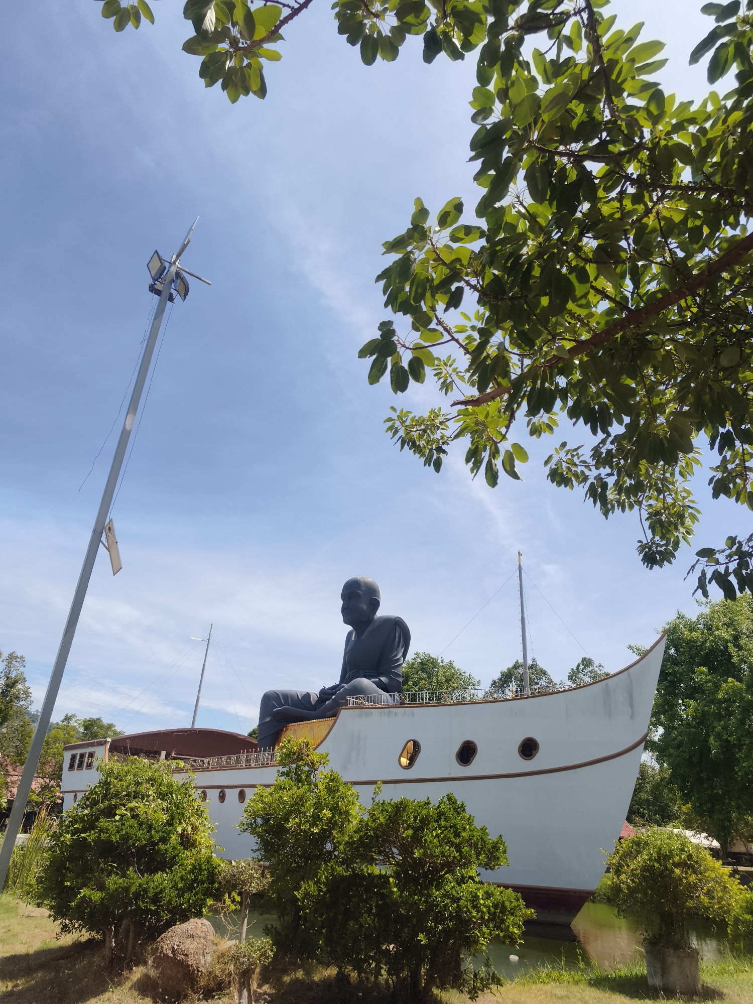 A low-angle, medium shot captures a large statue of a seated man on the deck of a white boat-shaped structure under a clear blue sky. The statue is dark, with the man's head bald and his eyes closed. He is seated with his legs crossed, wearing robes. The boat structure has a tan railing and several oval windows with a brown trim. Lush green trees and foliage frame the top and bottom of the image, with some branches reaching into the frame from the top. A tall, slender pole with lights and wires extends from the left side of the frame, piercing the sky. The boat structure appears to be situated in or near water, with reflections visible at the bottom. The scene is bathed in bright daylight. This statue is located at Wat Phra Yai, also known as the Big Buddha Temple, in the Ko Samui District of Thailand.