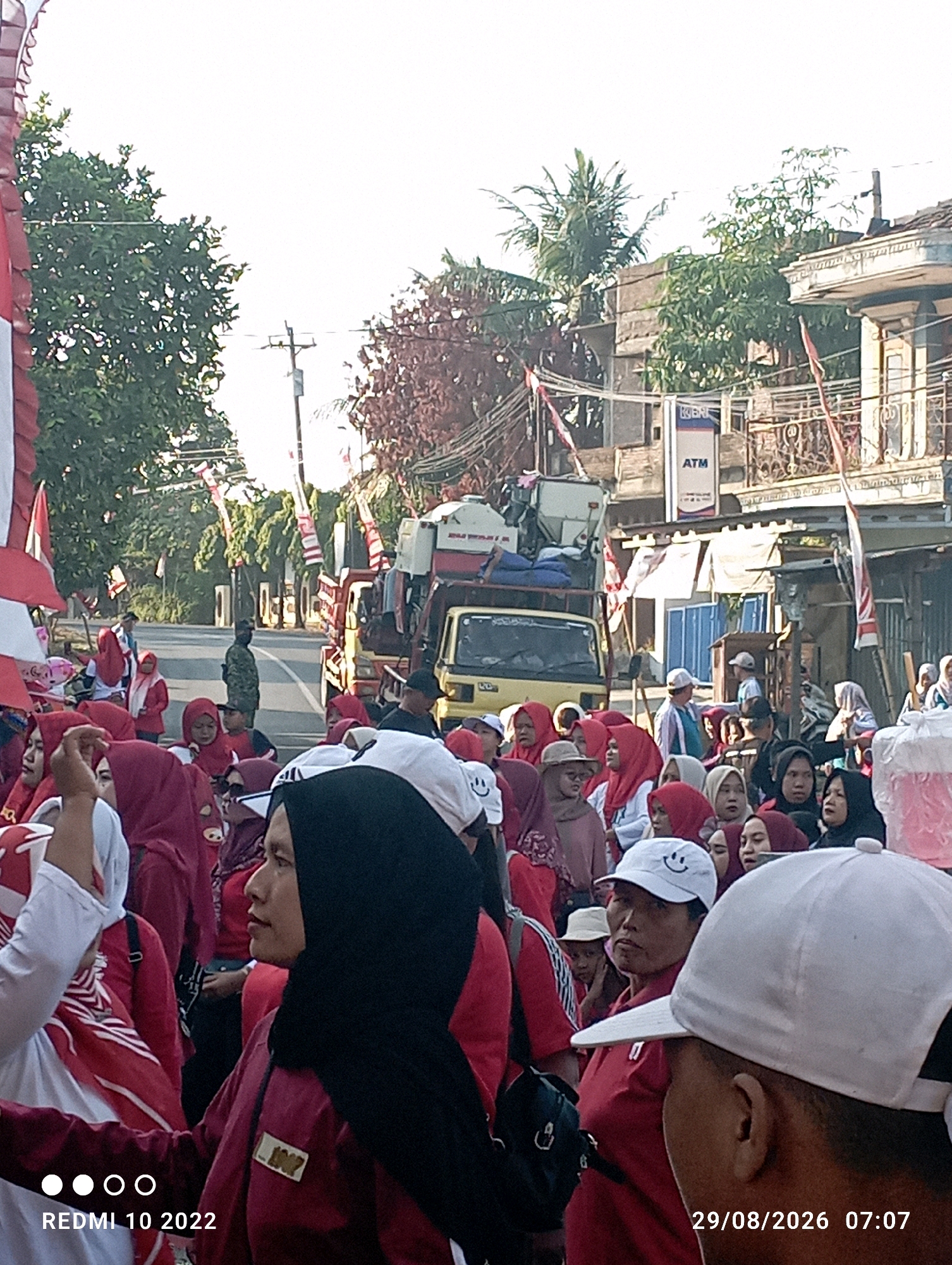 This image captures a street scene in Dukuhseti, Indonesia, on August 29, 2026, at 07:07. A large crowd of people, predominantly women and children wearing red and white attire, are gathered on the side of a road. Many are wearing hijabs, and some have white hats. In the background, a yellow truck with a white cargo box is visible, suggesting some form of procession or event is taking place. Buildings, trees, and utility poles line the street. A uniformed individual, possibly a soldier or security personnel, stands near the center of the road. Flags, likely Indonesian national flags, are displayed along the street and on some vehicles. The overall atmosphere appears festive or celebratory.