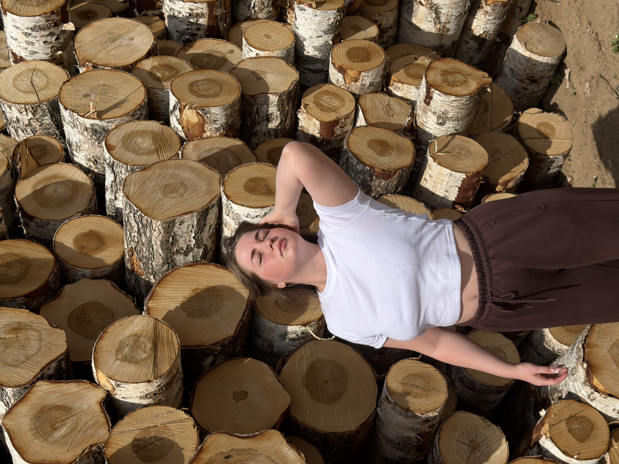 A high-angle shot shows a woman lying on a pile of birch logs in what appears to be an outdoor setting. She is positioned diagonally across the frame, her head toward the left and feet extending toward the right.
The woman is fair-skinned with light brown hair. She is wearing a short-sleeved white T-shirt that exposes a small portion of her stomach and dark brown sweatpants. Her right arm is resting by her side, while her left arm is bent with her hand near her head. Her eyes are closed, suggesting she is resting or asleep.
The surface beneath the woman is composed of numerous birch logs stacked closely together. The logs are light in color, with distinctive white bark and visible growth rings on the cut ends.
The lighting is bright and appears to be natural sunlight.
There is no indication of the location being specifically Ural'skoe, Russia, from the visual evidence provided. There are no city-specific landmarks or identifiers present. The scene appears to be in a wooded area or storage area for firewood.